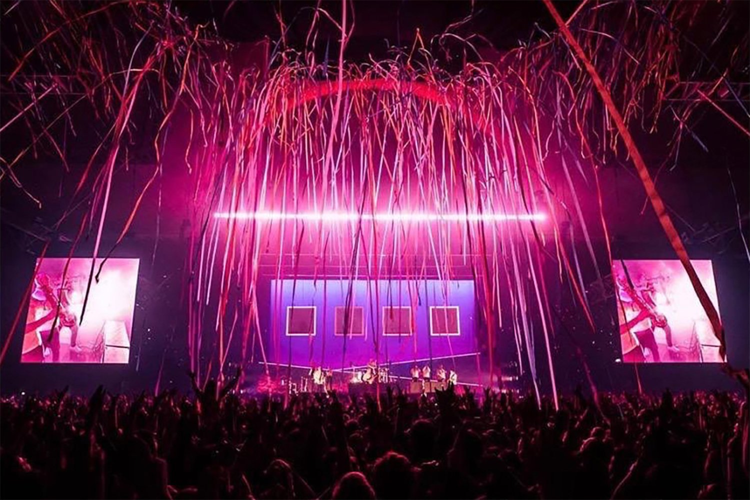 Pink and purple concert stage with streamers and screens, audience silhouette in front, performers on stage, vibrant lighting and decorations.