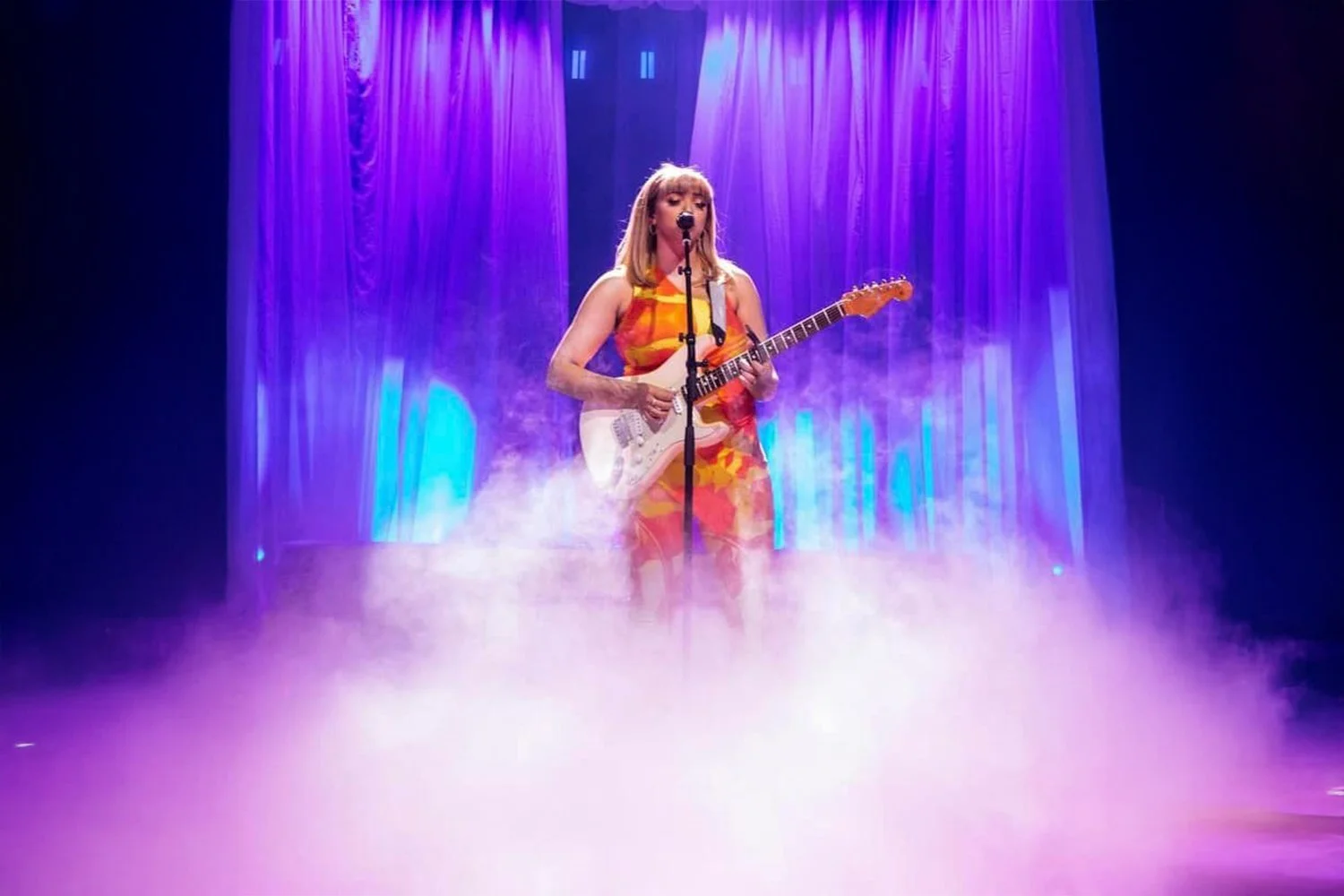 A woman performing on stage with an electric guitar, surrounded by purple and blue stage lighting and fog
