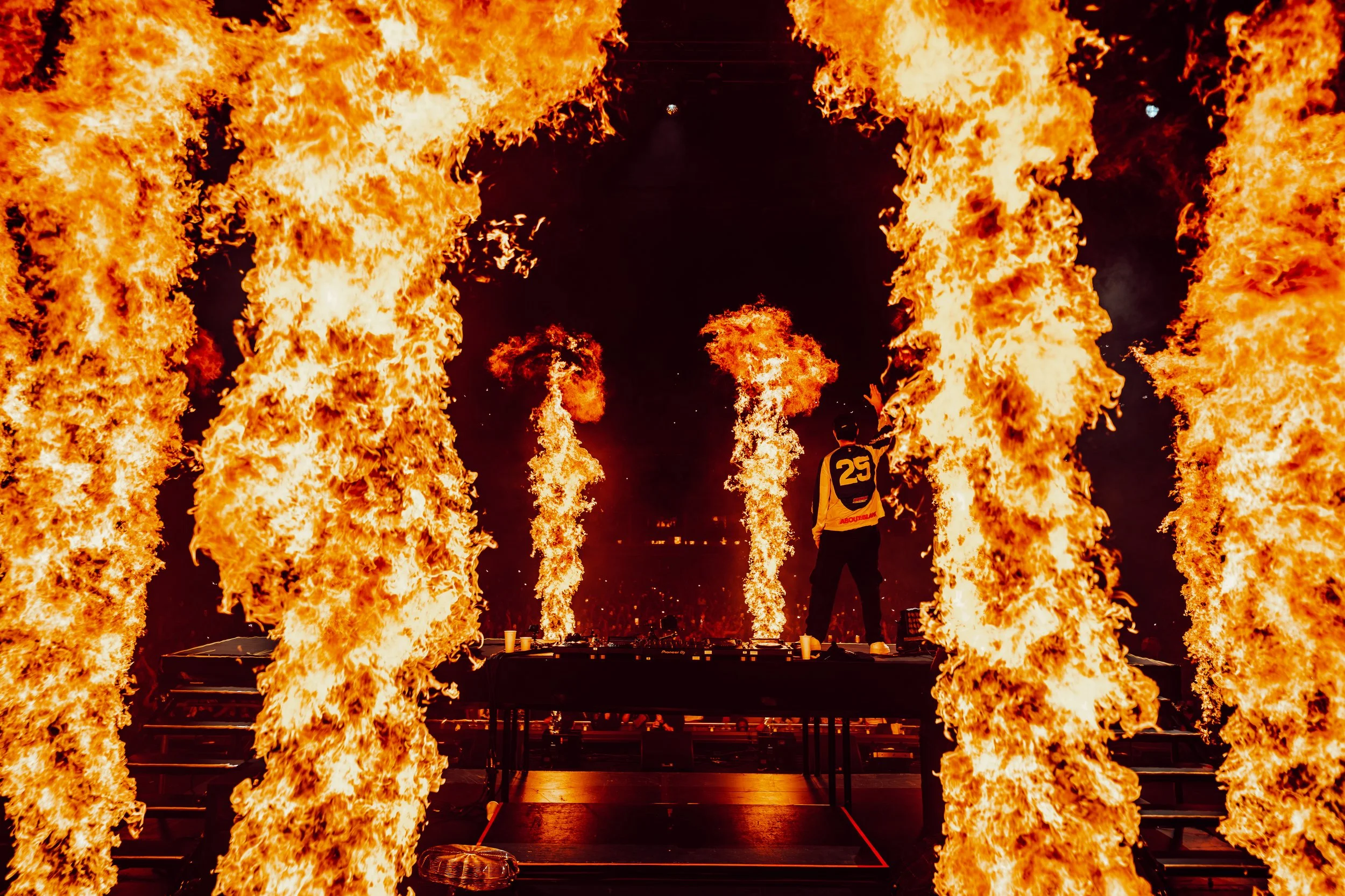 A person in a yellow jacket with the number 25 on the back stands amidst a large fire showcase, with tall flames on either side of the scene and some sparks flying in the air.