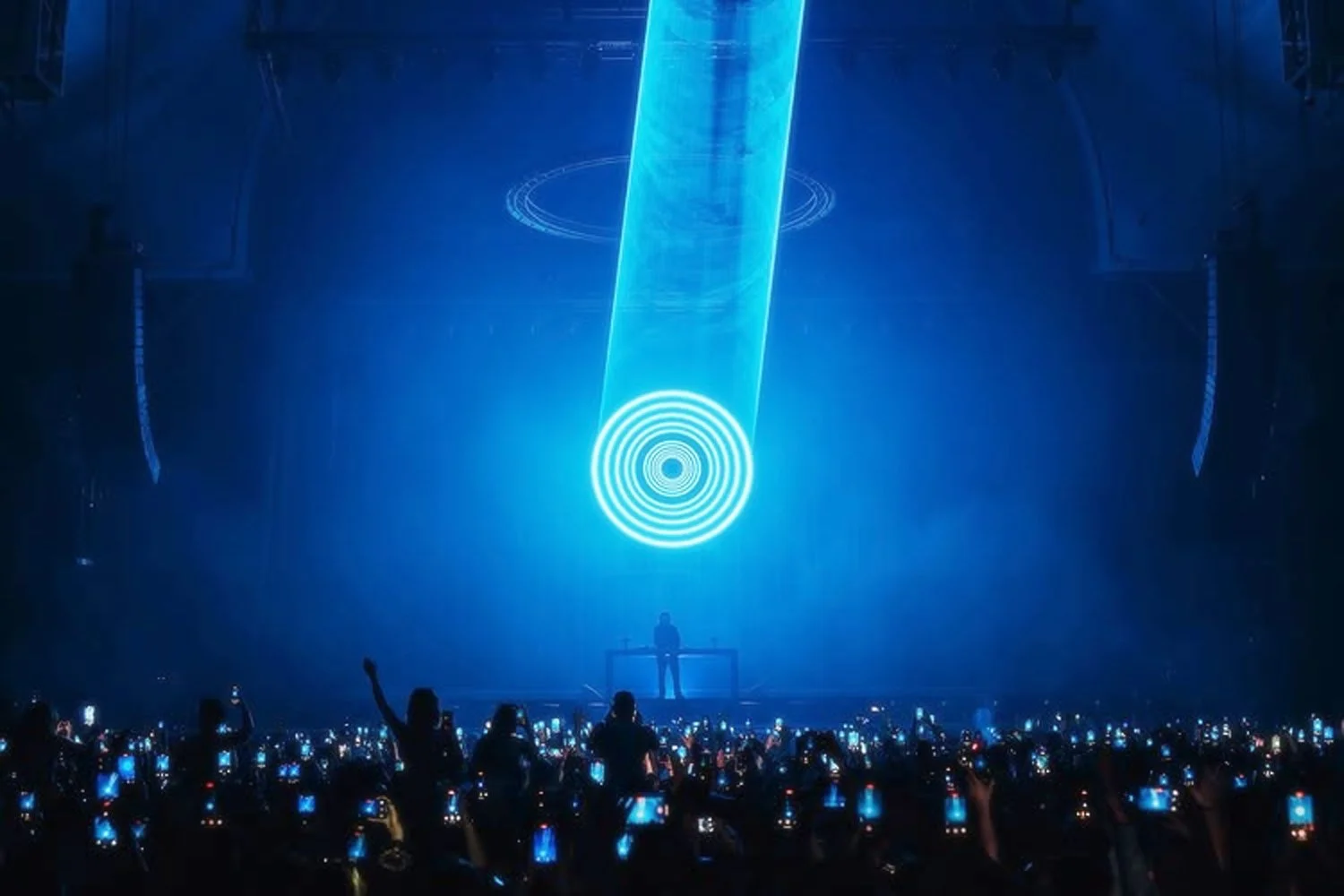 A concert with a performer on stage, illuminated by a blue laser beam. The crowd holds up their phones, creating a sea of lights.