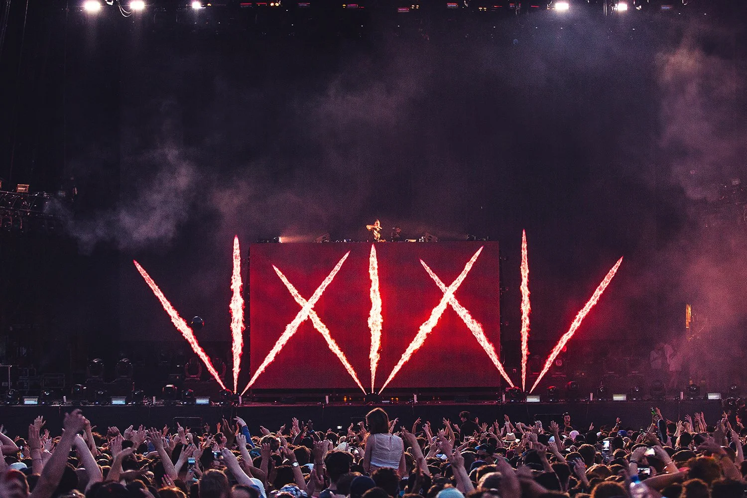 A live concert with a large crowd, a stage with a DJ behind a screen, and vibrant red flame effects on the screen forming two X's and the number 777.