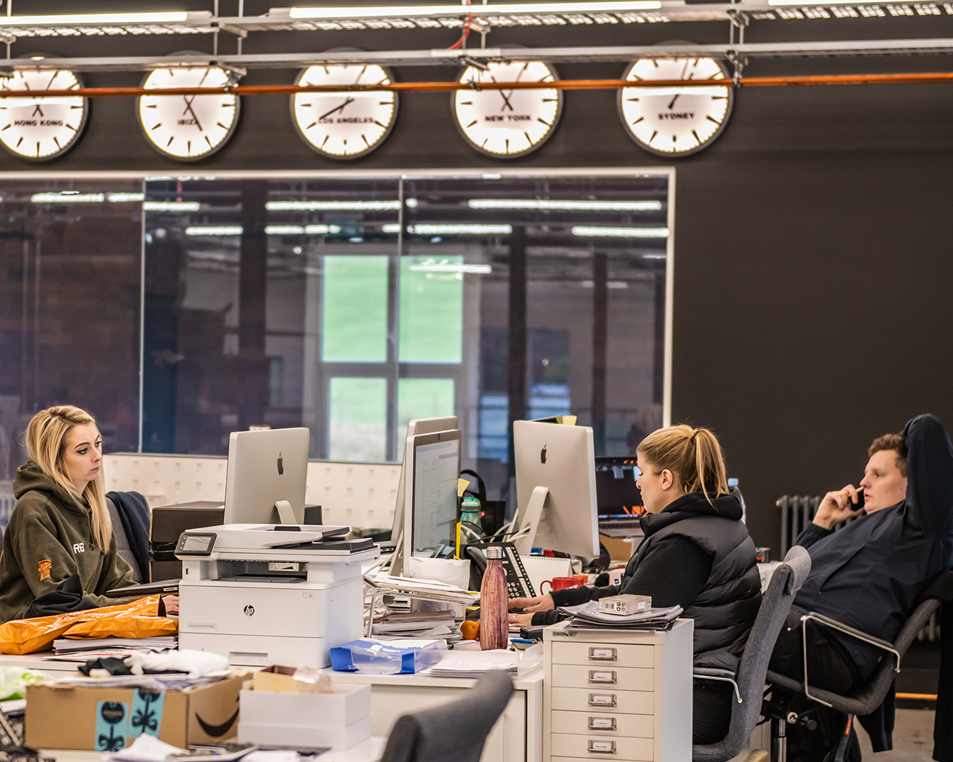 An office environment with four people working at desks with computers, paperwork, and office supplies. There are four clocks on the wall showing different world times, and a large window in the background.