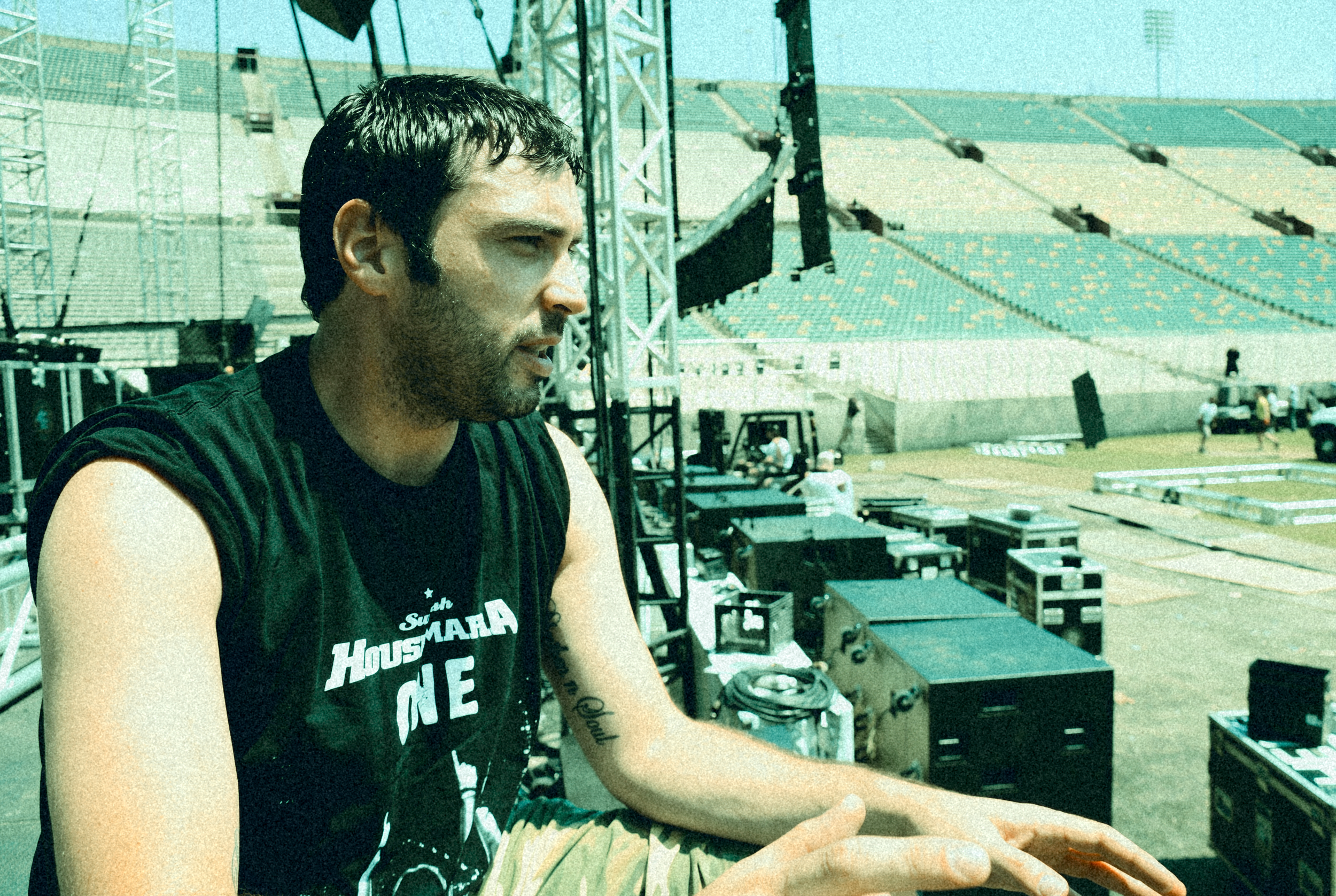 A man with dark hair and stubble, wearing a black sleeveless shirt, sitting at a stage setup in a large outdoor stadium. The background shows empty stadium seats, stage equipment like speakers, lighting, and scaffolding.