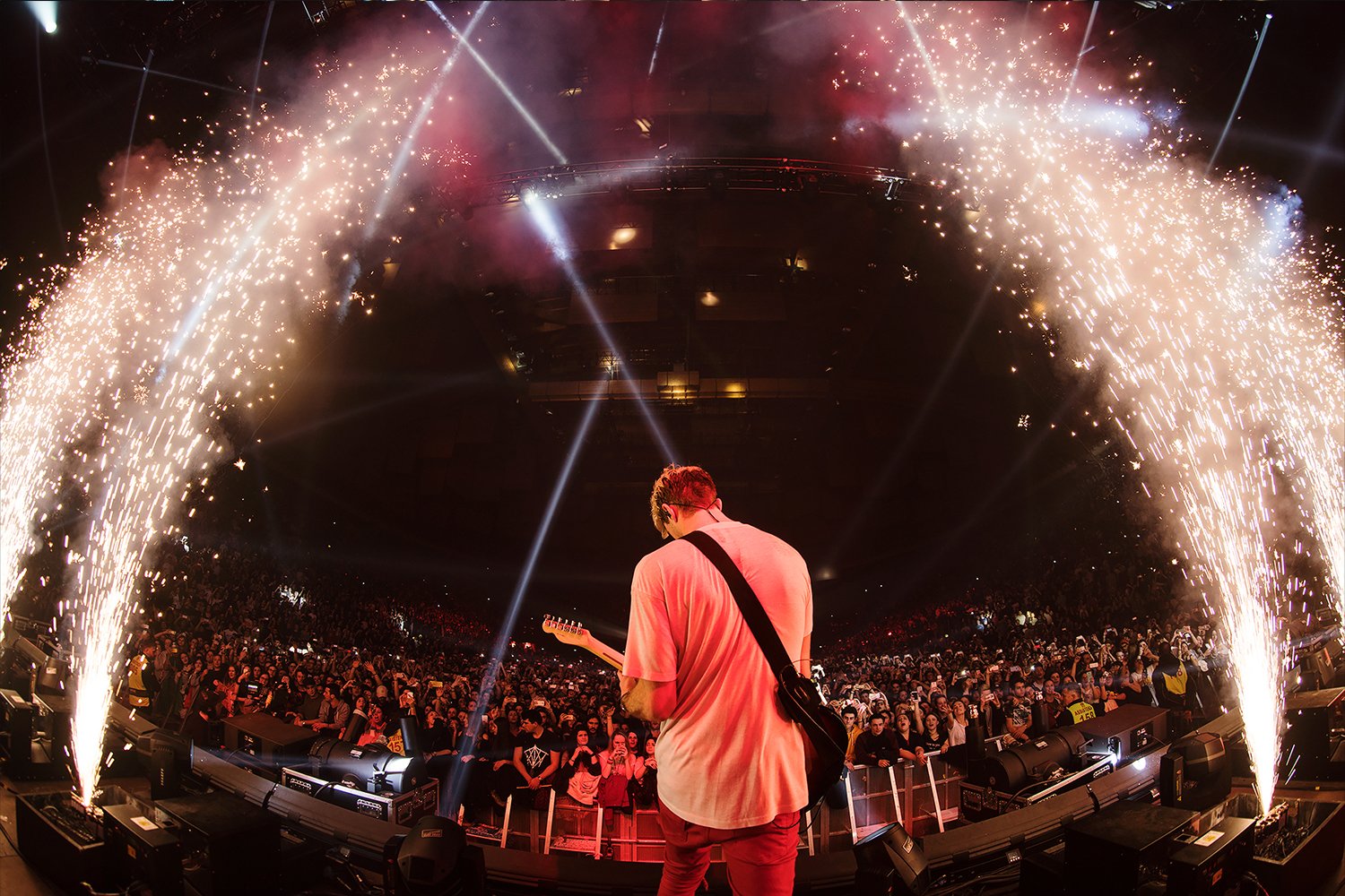 A musician performing with a guitar on stage at a concert with pyrotechnics and bright lights, and a large crowd of fans in the background.