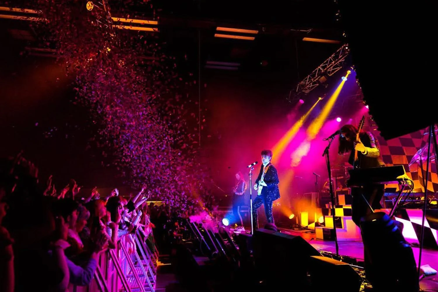 Concert scene with a band performing on stage, colorful stage lighting, and confetti in the air. Audience members are cheering near the barricades.