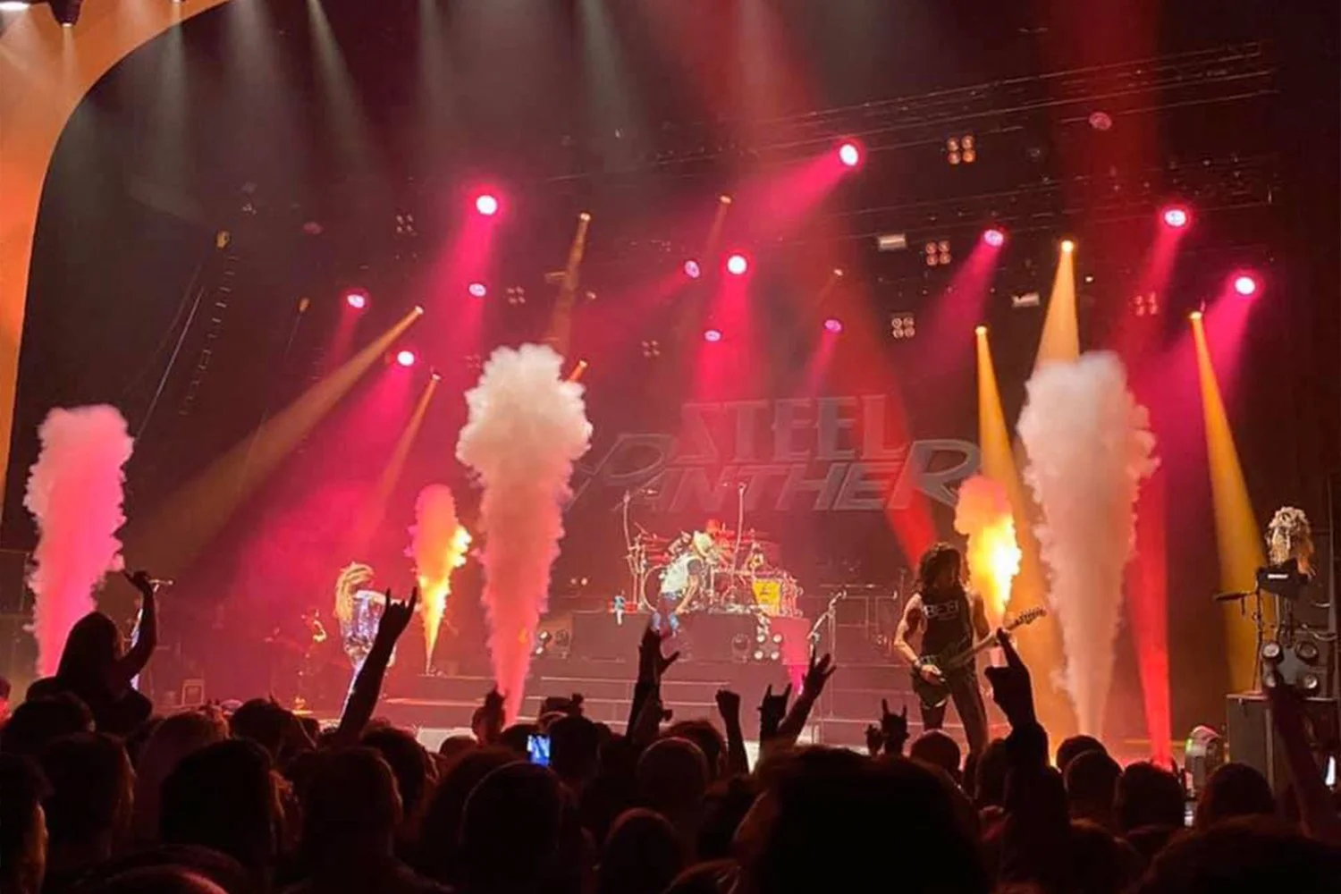 A live concert on stage with colorful lighting, smoke effects, and a crowd cheering. A guitarist is playing near the front of the stage, and the band name 'Steel Panther' is displayed in the background.