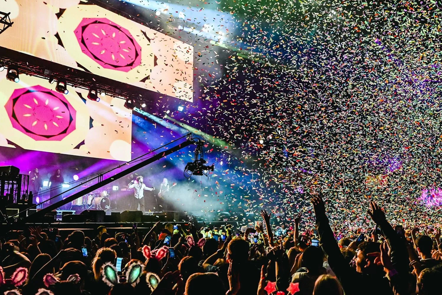 Concert scene with a large crowd, colorful confetti, bright stage lights, and a band performing on stage.