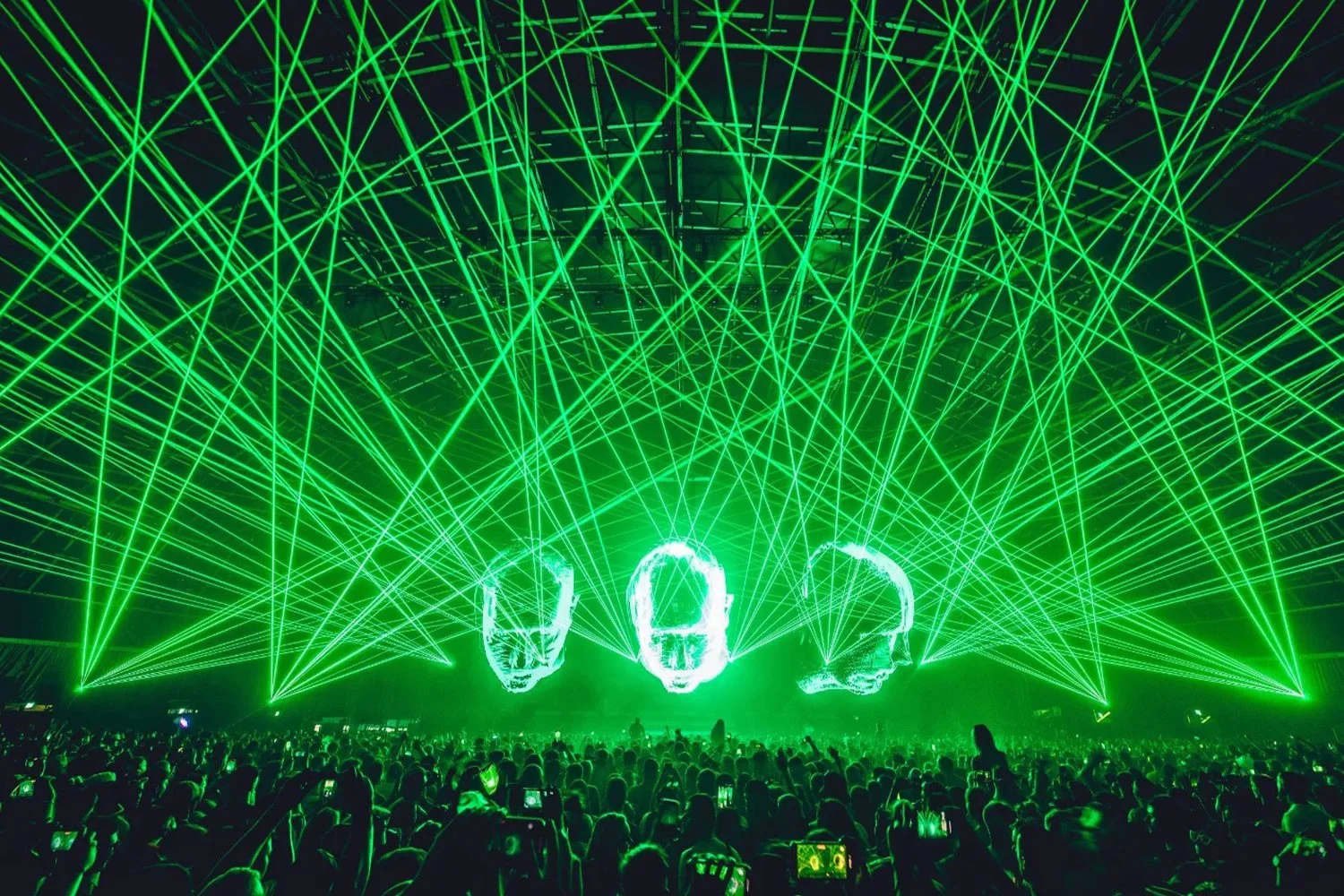 A concert featuring three large skull images displayed on screens with a green laser light show overhead and a large crowd of people watching.