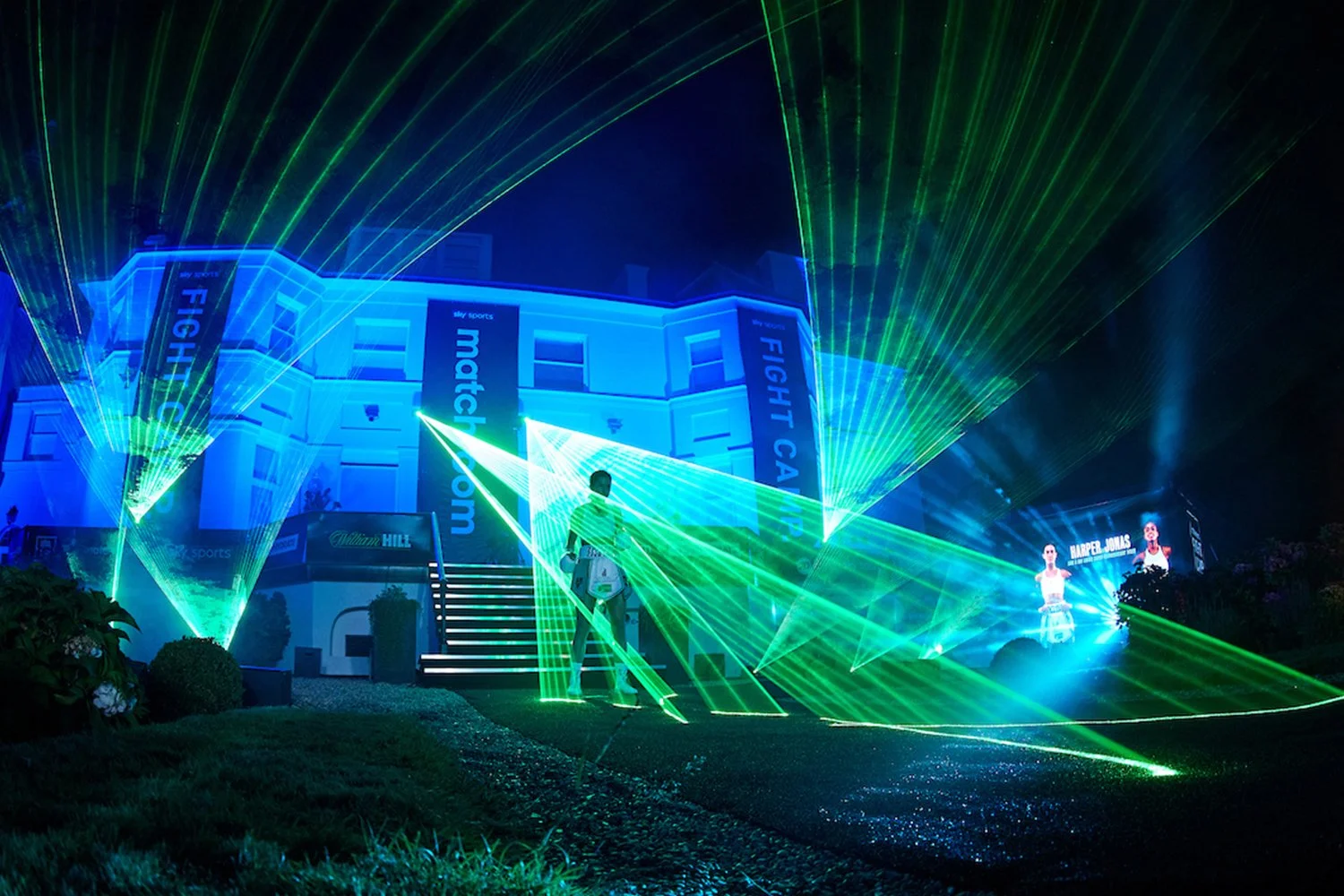Nighttime scene with green laser lights projected onto a two-story house, with two individuals standing in the foreground, and large screens displaying tennis players and name Harper Jones in the background.