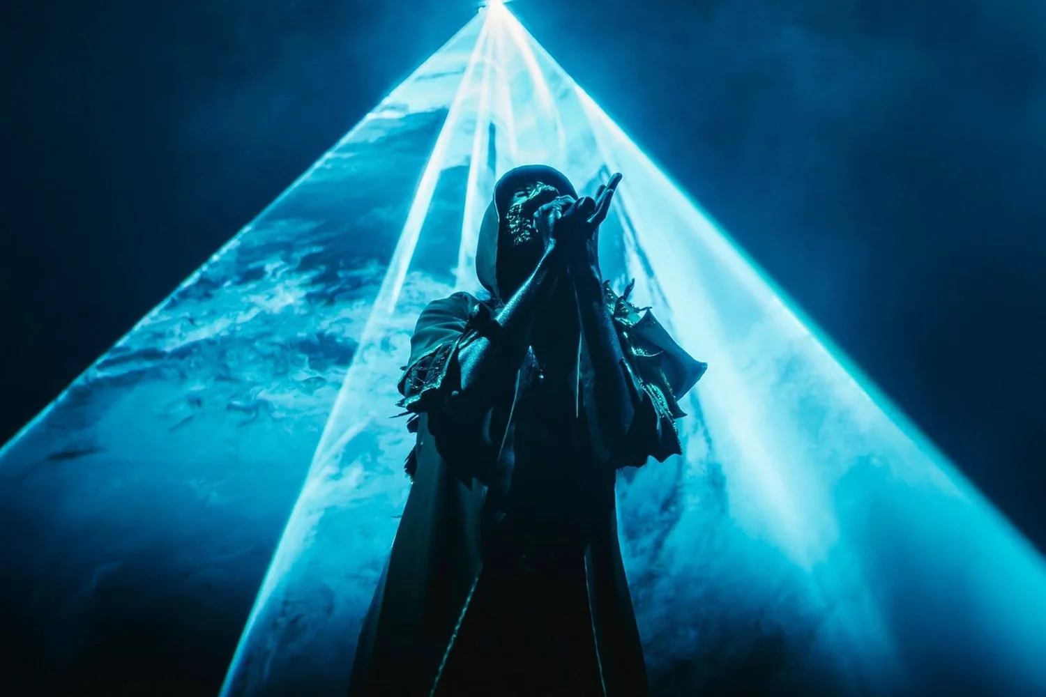 A person in dark clothing and a mask is singing or speaking into a microphone on stage with blue laser lights and a background that resembles sky and clouds.