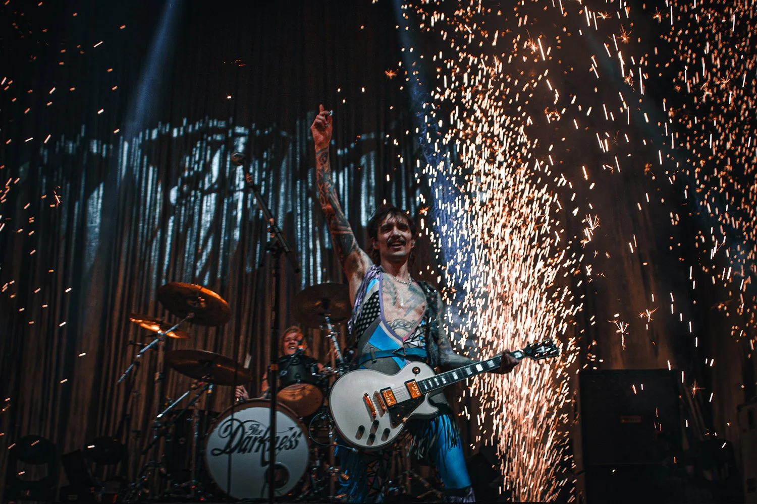 Musician on stage with guitar performing during fireworks display, with drummer behind, at a concert.