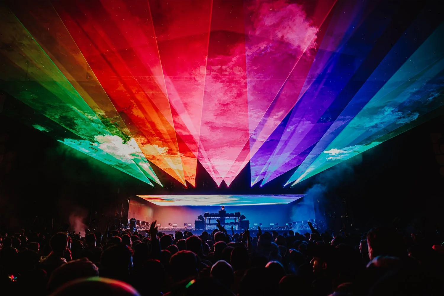 A concert with a DJ performing on stage, colorful laser lights projecting sky and cloud images in rainbow colors, and a large crowd in the foreground.