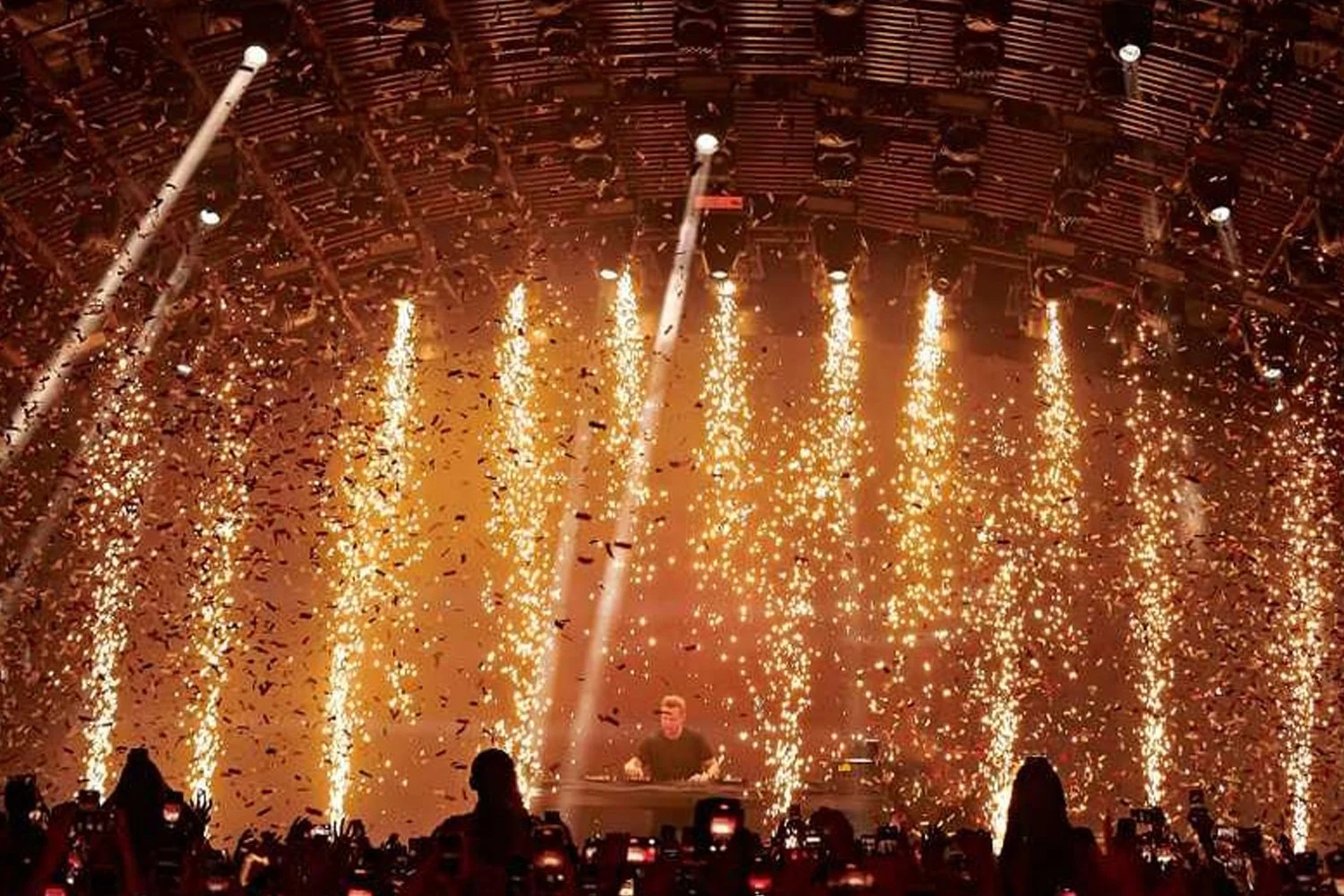 A DJ performs on stage with a background of vertical firework or spark effects and falling confetti, with audience members taking photos.