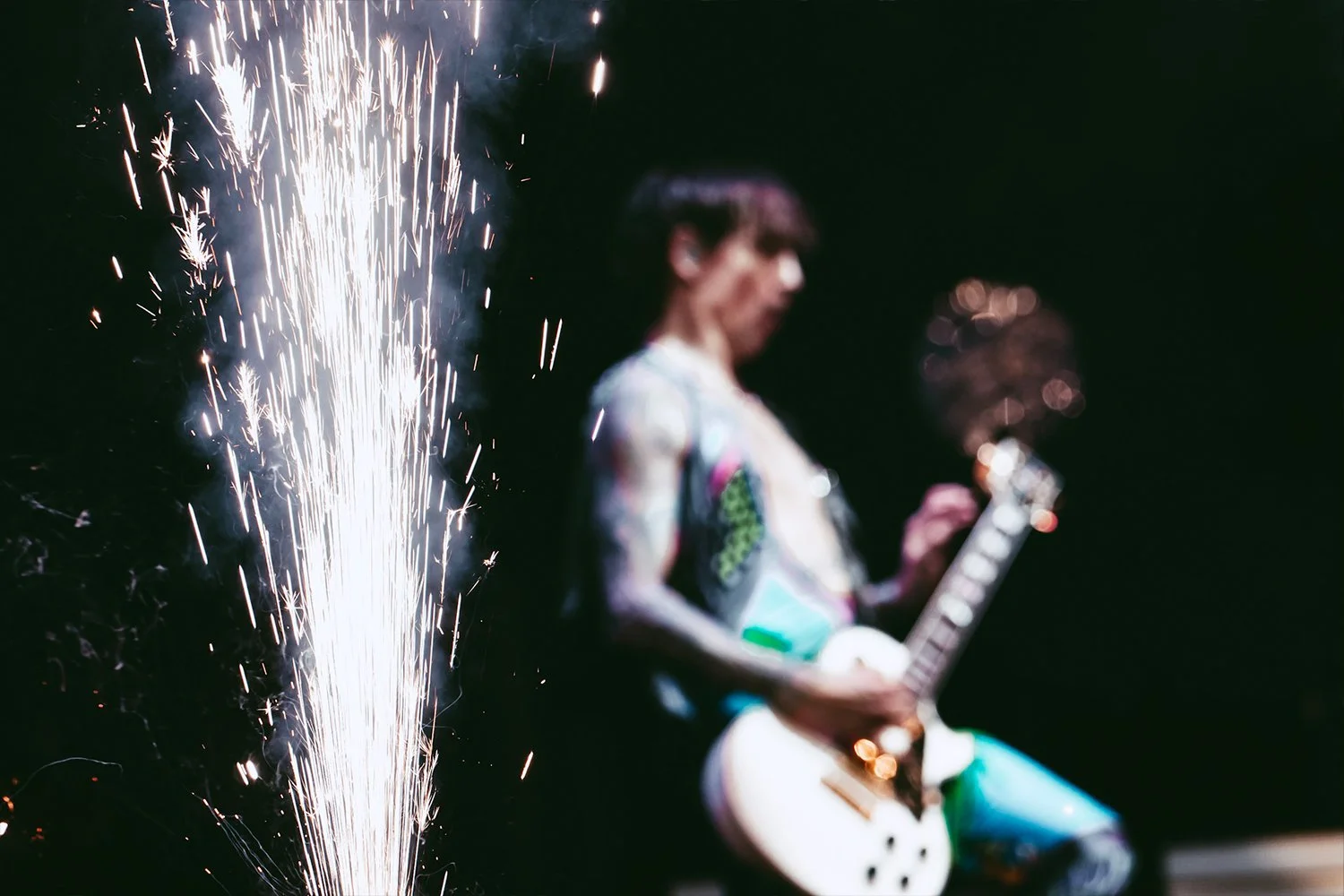 Blurred image of a musician playing an electric guitar with bright sparks or fireworks in the foreground, set against a dark background.