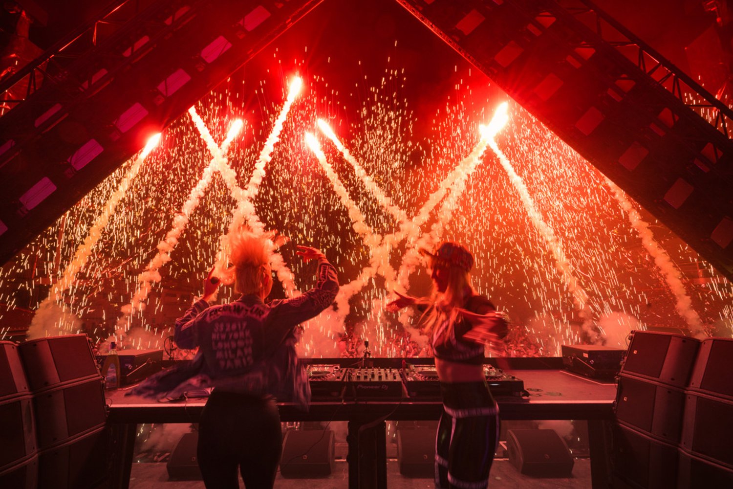 Two female DJs playing music at a nightclub with fireworks and sparks exploding in the background.