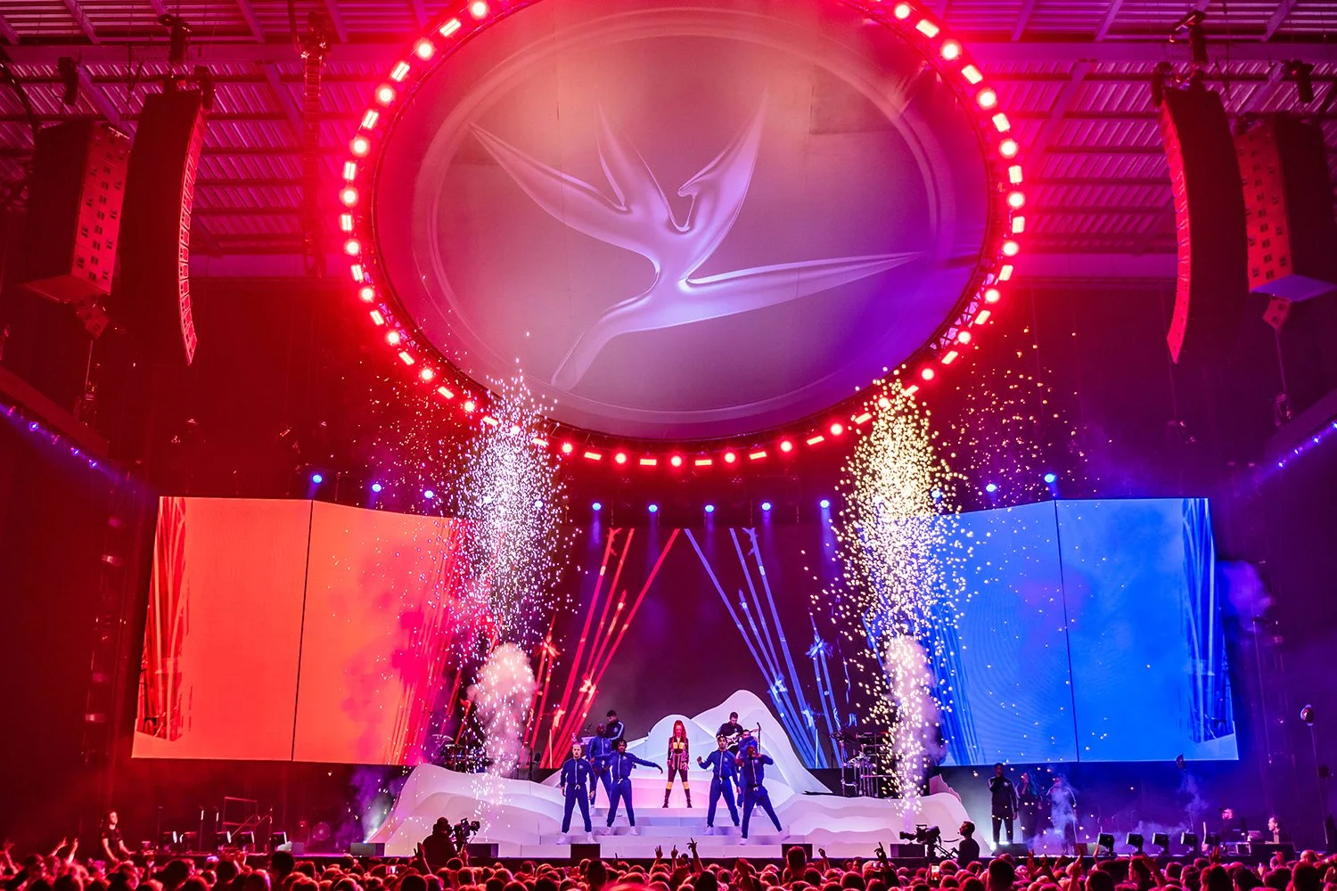 Concert stage with performers, colorful lighting, and fireworks display.