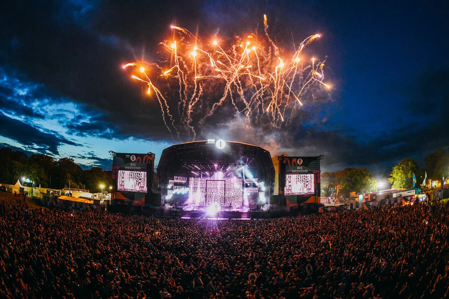 A large outdoor concert stage illuminated with purple and white lights, with a crowd of thousands watching, and fireworks exploding in the night sky above.