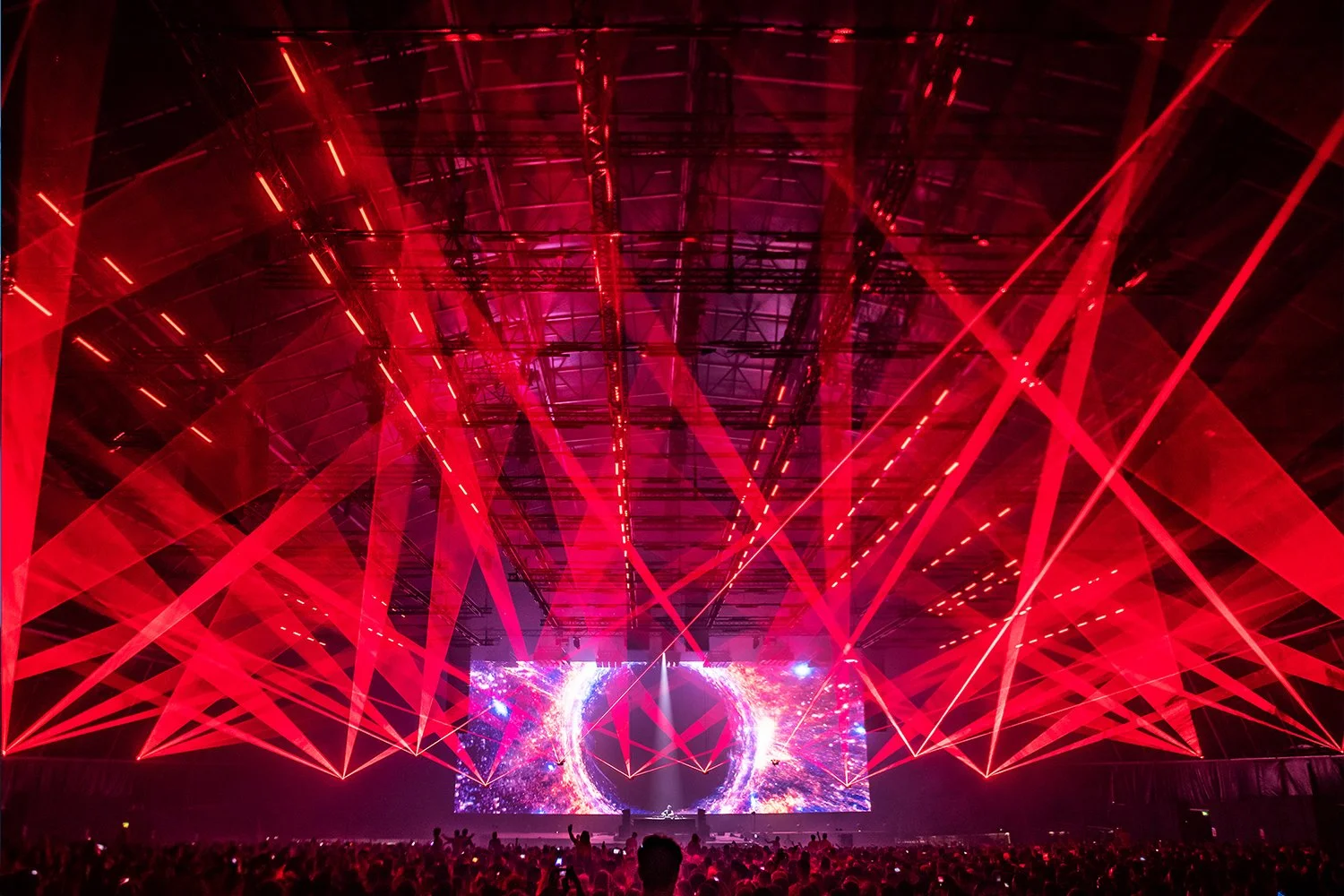 Concert with red laser lights and a large screen displaying colorful visuals, crowd in the foreground
