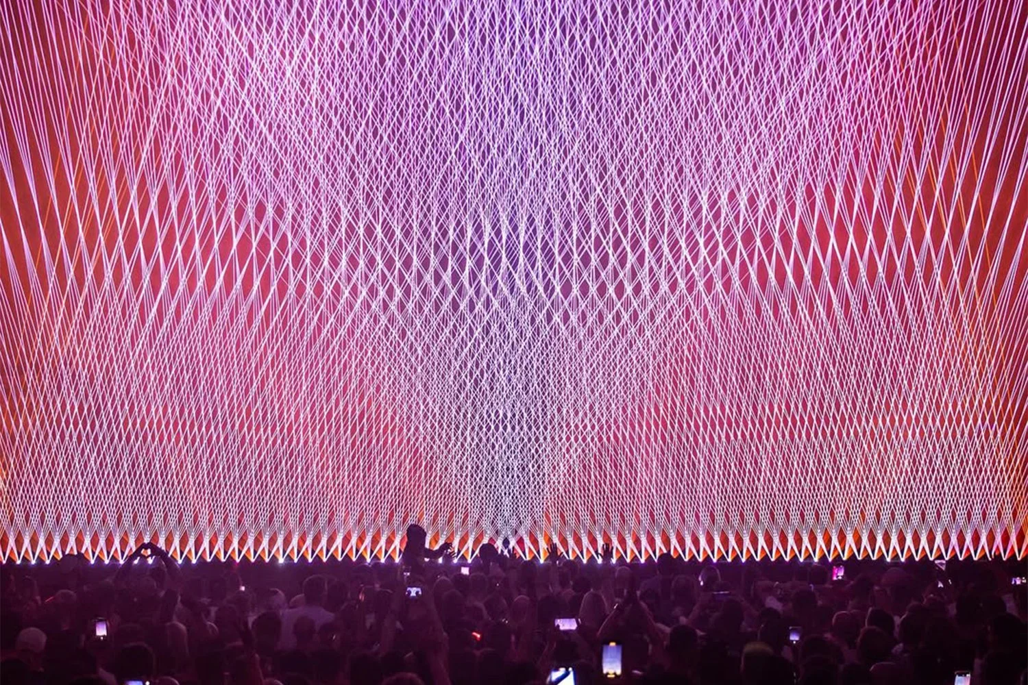 Large crowd of people watching a vibrant light and laser show with purple, pink, and orange colors projecting intricate geometric patterns on a large screen.