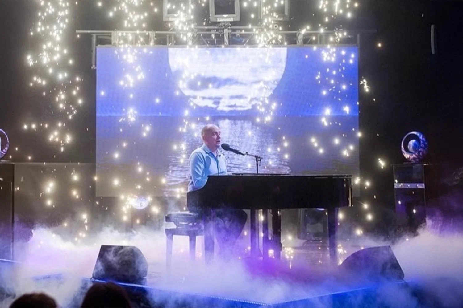 A man playing a grand piano and singing into a microphone on a stage with bright lights and a large screen displaying a moonlit water scene behind him. There is fog on the stage floor, and decorative elements with glowing orbs are visible.