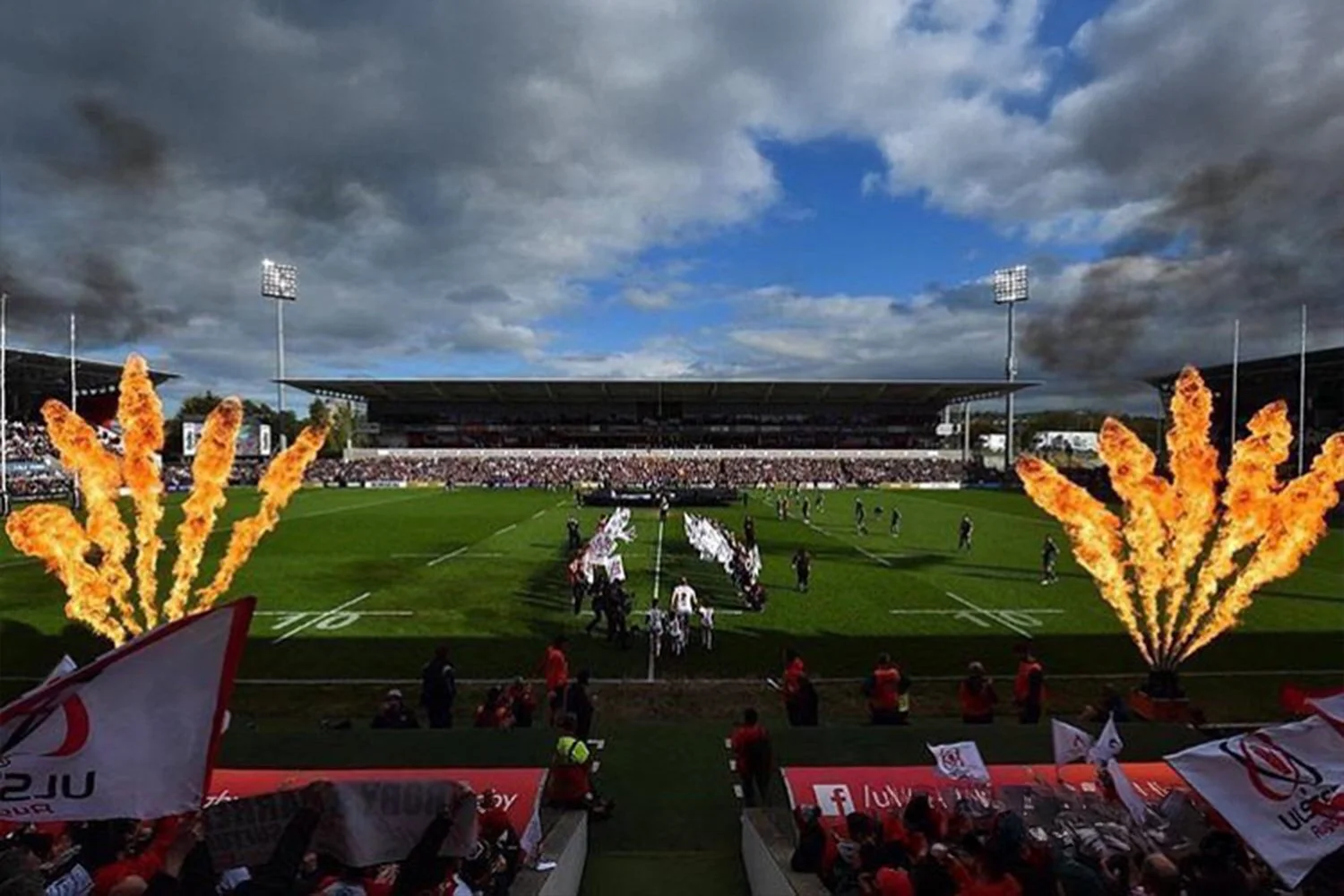A sports stadium filled with spectators during a game, with players on the field and explosions of fire on either side of the entrance, under a partly cloudy sky.
