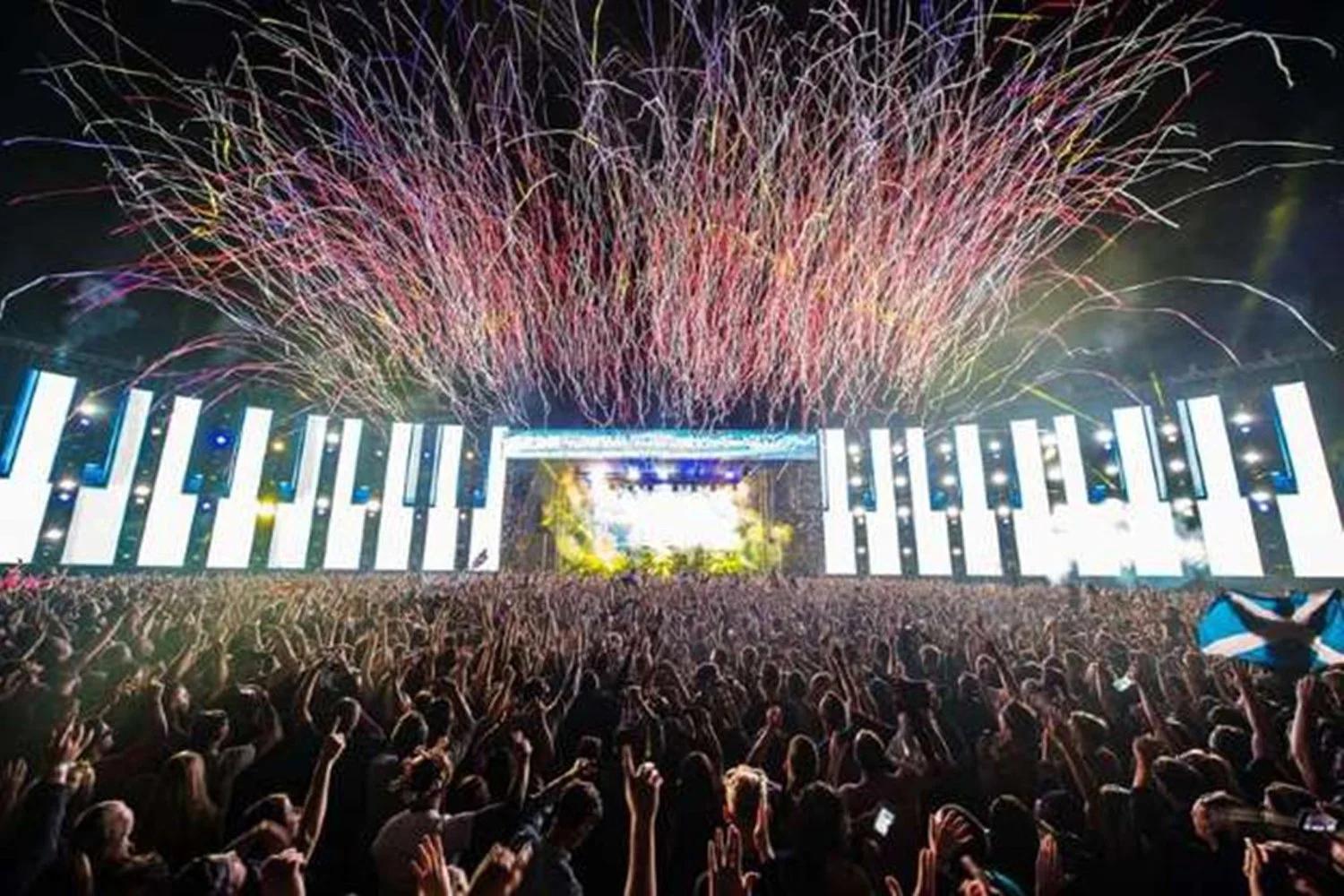 A large crowd at a concert or festival with fireworks and colorful lights above the stage.