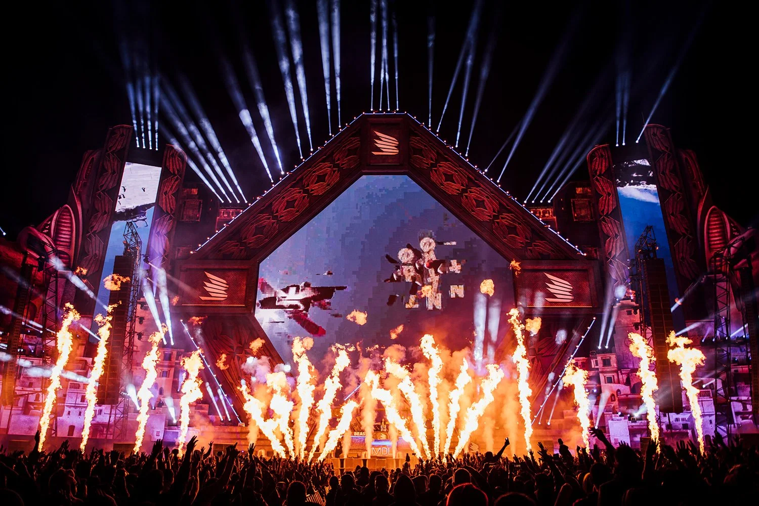A concert stage with pyrotechnics, bright lights, and large screens displaying visuals, with a crowd of people cheering and raising their hands.