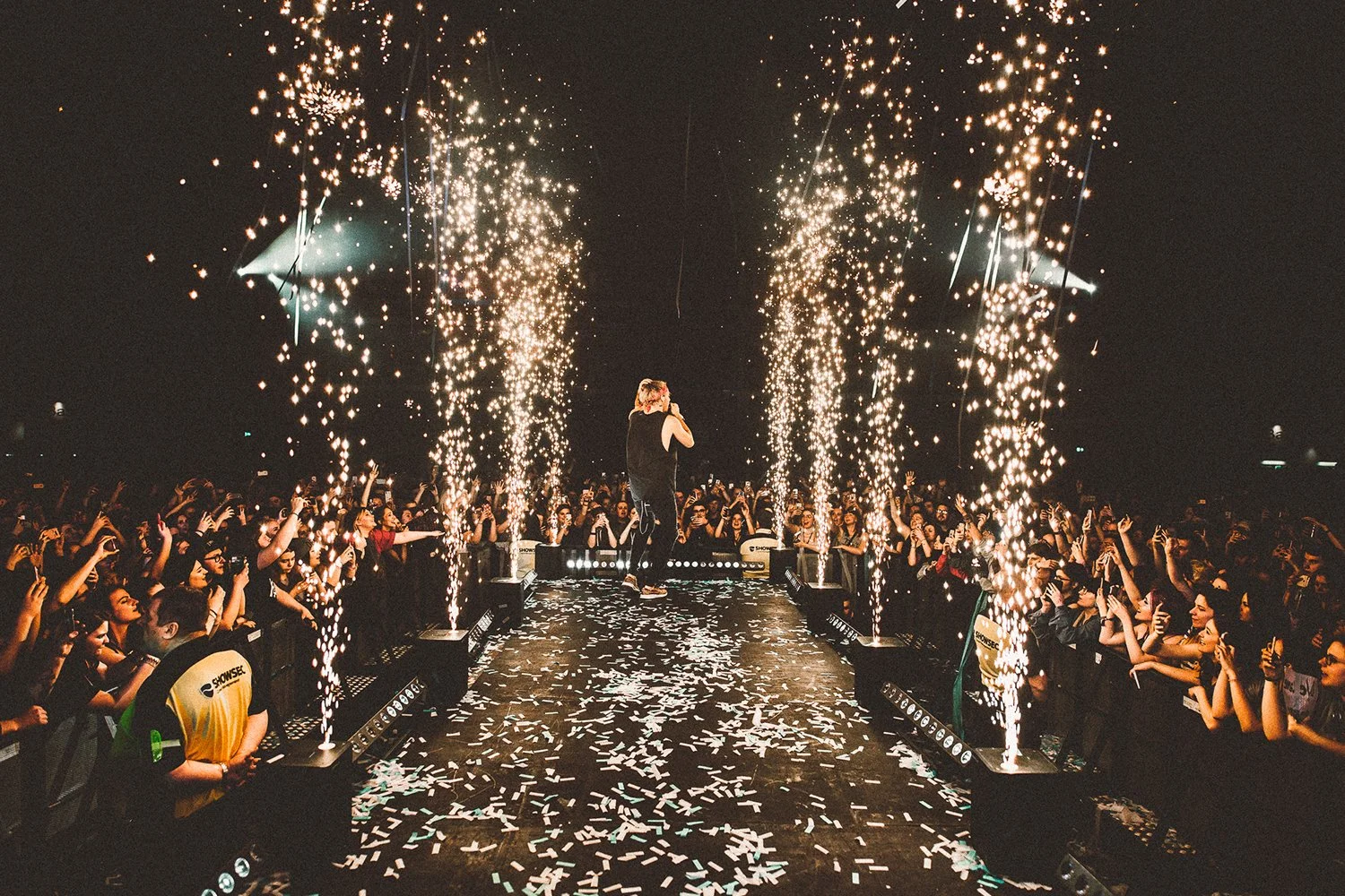 A concert stage with a performer singing or speaking into a microphone, surrounded by audience members cheering and raising their hands. Fireworks and confetti are being released above the stage, creating a celebratory atmosphere.