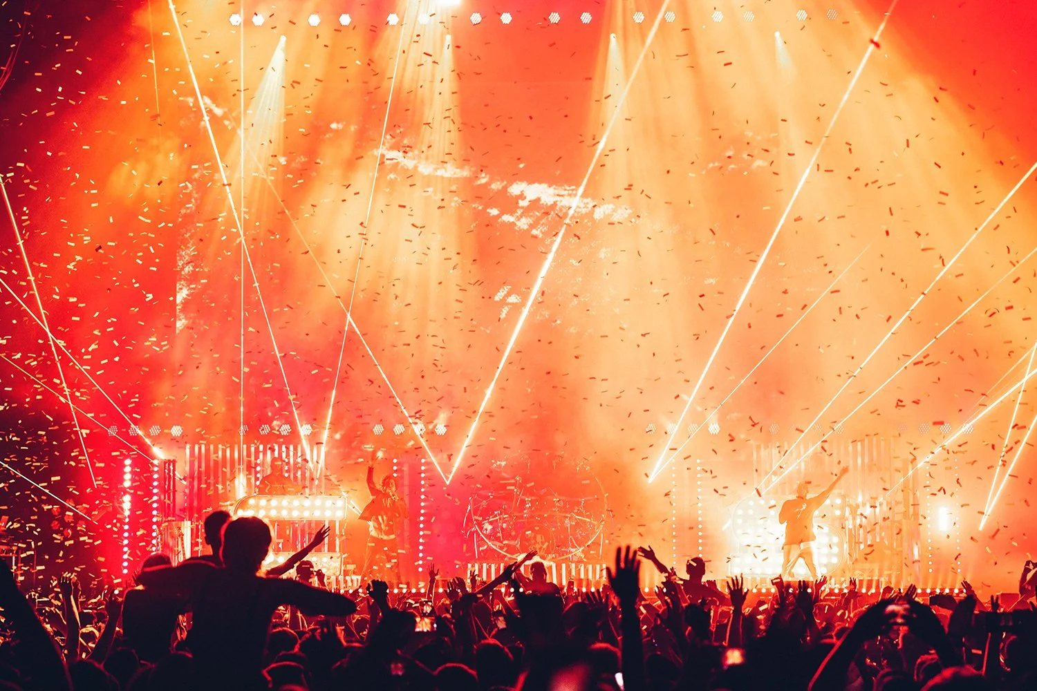 A live concert with bright orange and yellow stage lights, confetti, and laser beams, with a crowd of people watching and raising their hands.