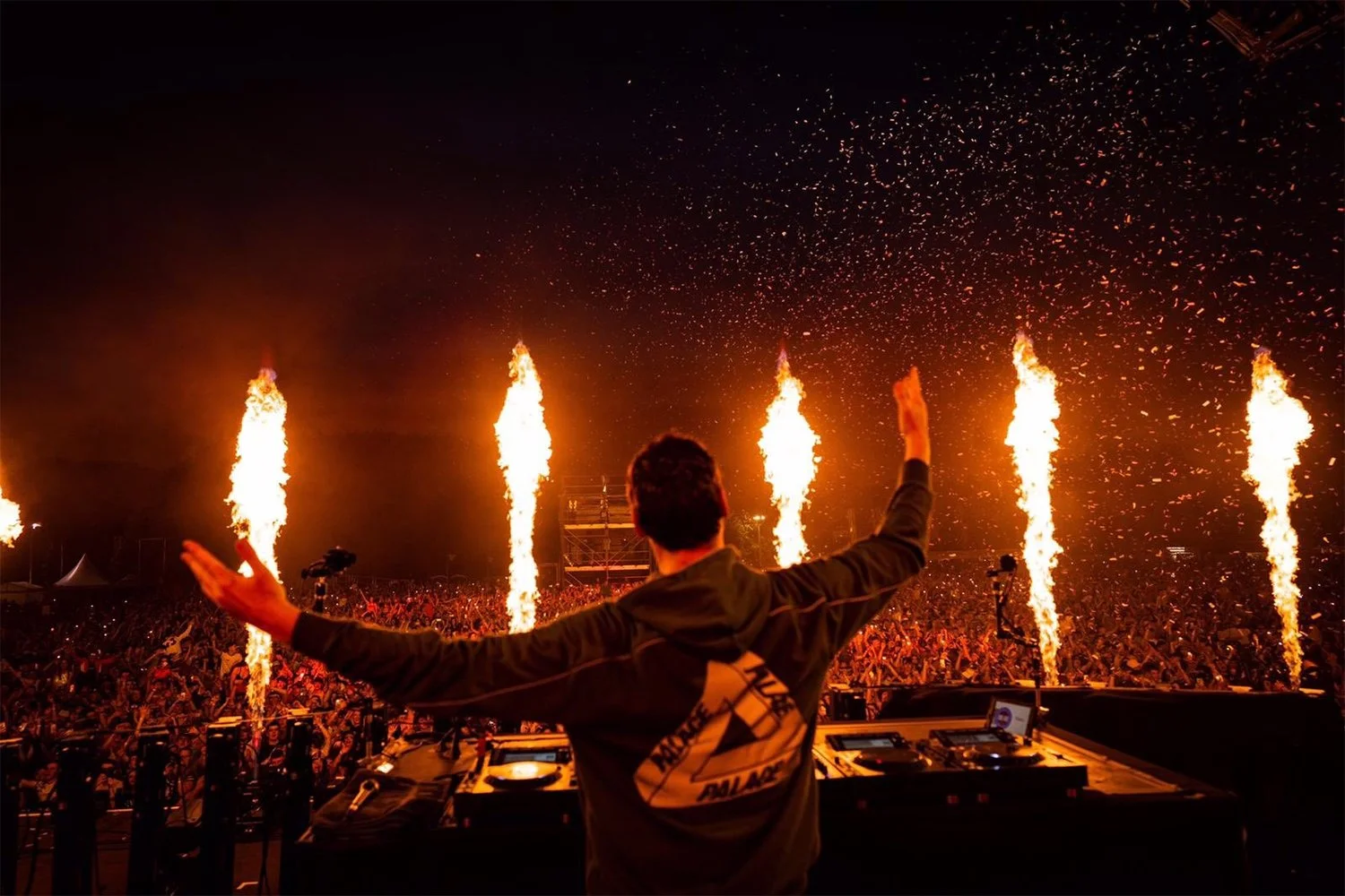 DJ performing at a concert with fire effects and pyrotechnics, and a large crowd in the background.