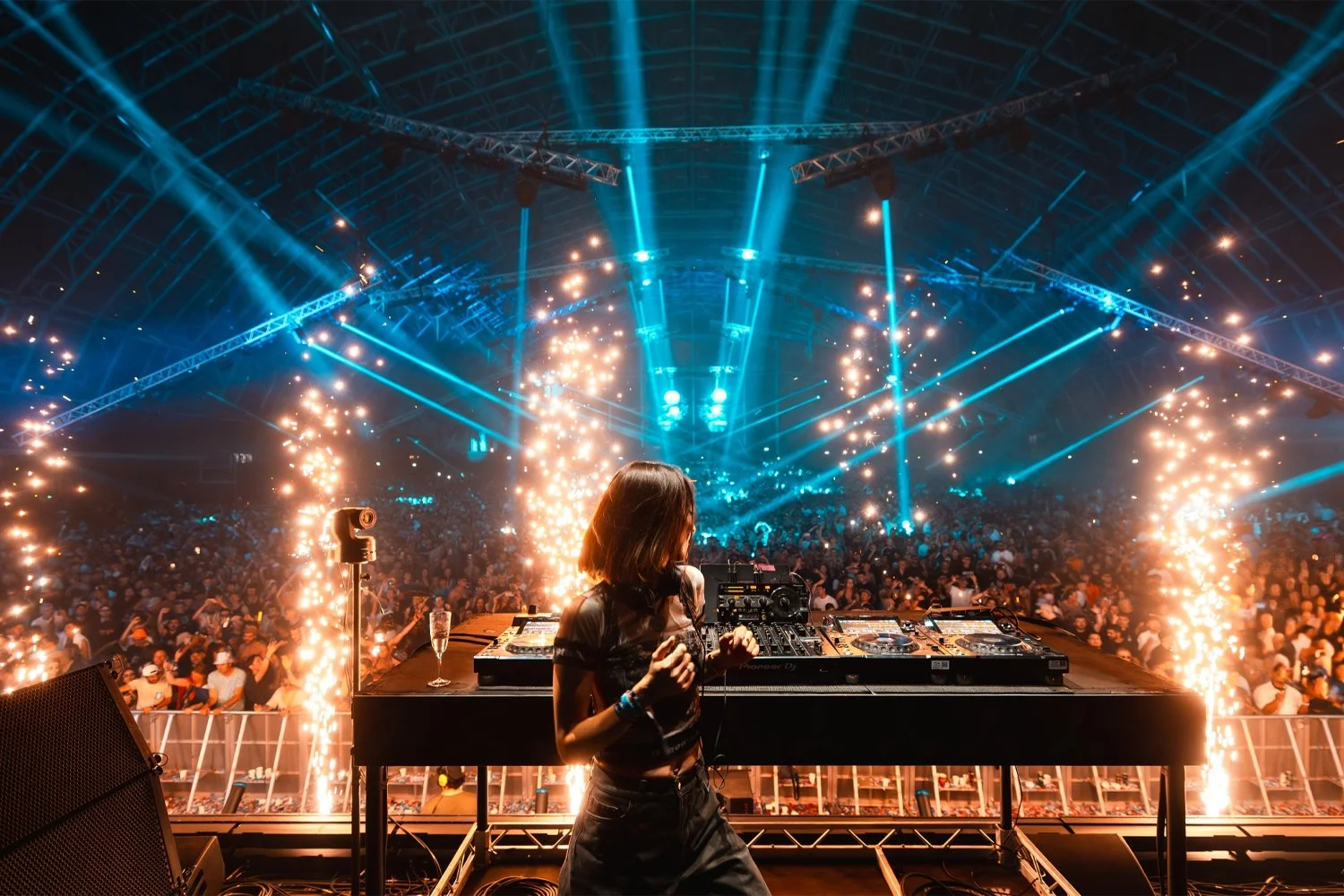 Female DJ performing on stage at a concert with bright blue and orange lighting, pyrotechnics, and a large crowd in the background.