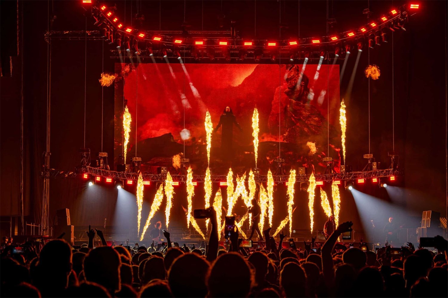 A concert stage with performers and pyrotechnics, featuring bright flames and a large screen displaying a fiery landscape in the background. Audience silhouettes are visible in the foreground.