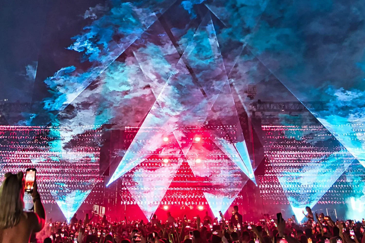 A large crowd at a concert or event with many people holding up phones. The stage features large screens and a light display showing red and blue lights and abstract patterns in the sky.