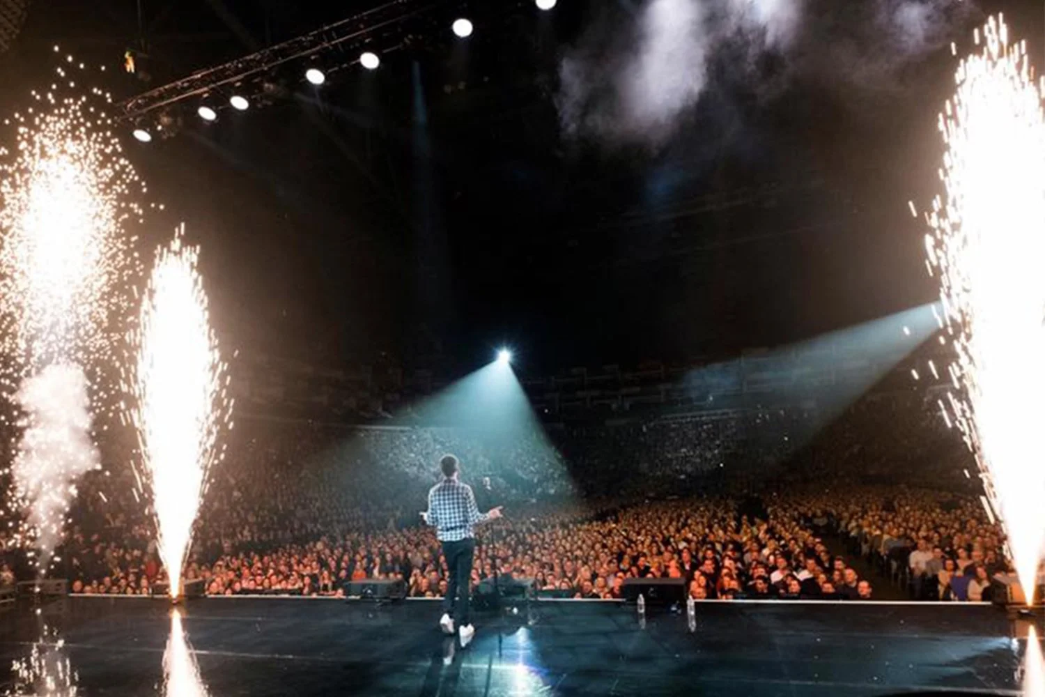 Performer on stage with fireworks and pyrotechnics, facing a large audience in a concert or event hall.
