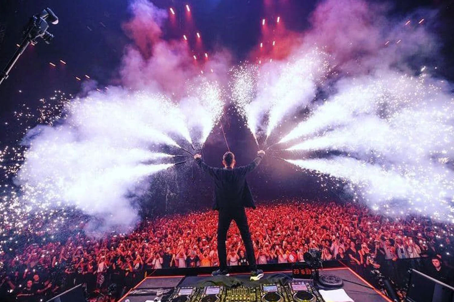A DJ performing on stage with pyrotechnics and fireworks, with a large crowd in front at a concert or music festival.