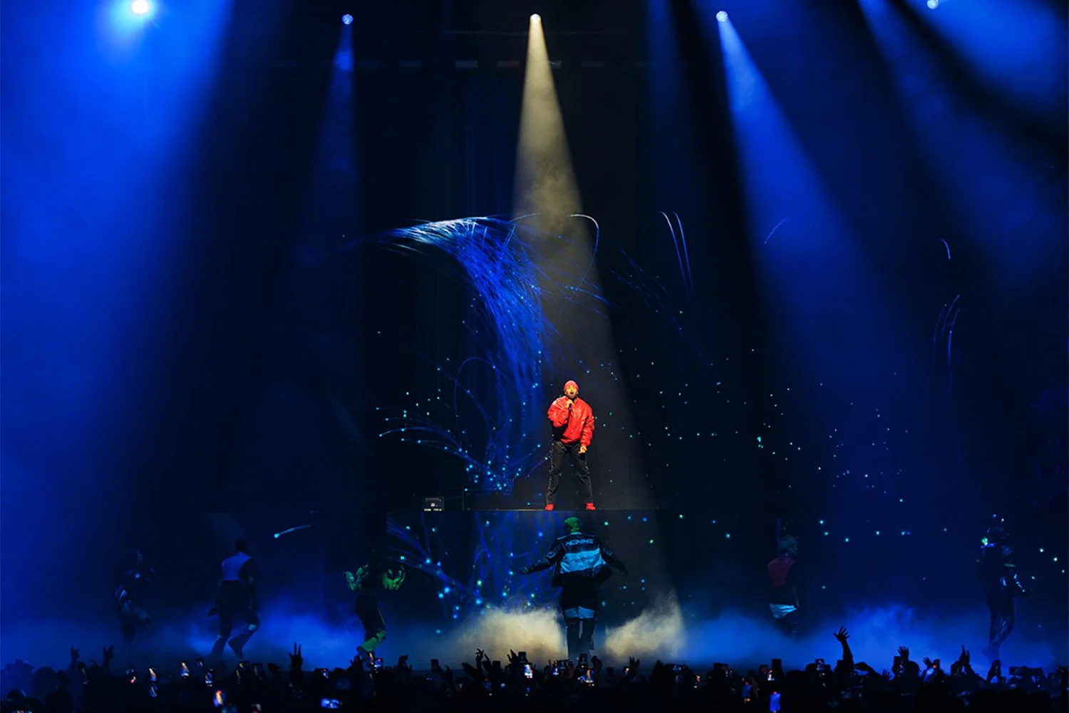 A performer on stage in a red jacket standing on a raised platform, illuminated by studio lighting, with dancers below and digital effects in the background.