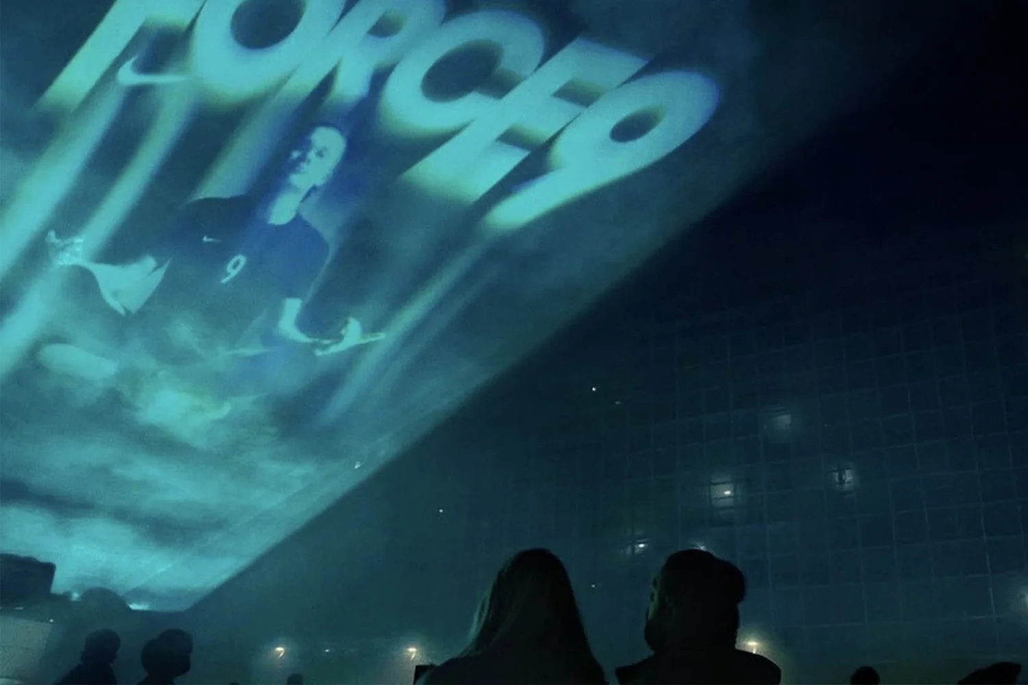 People watching a large projection of a soccer player in action on a wall.