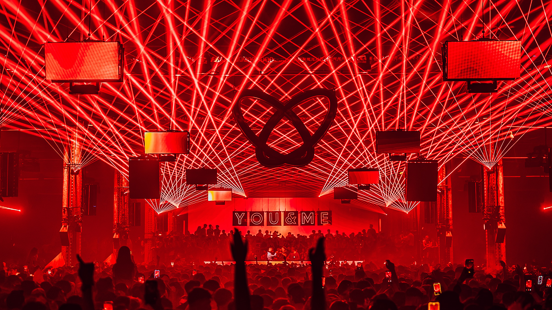 A concert or event stage illuminated with red laser lights and large screens. A prominent letter logo is displayed center stage, with the words 'YOU & ME' visible in the background. The crowd is visible at the bottom, with some people raising their hands.