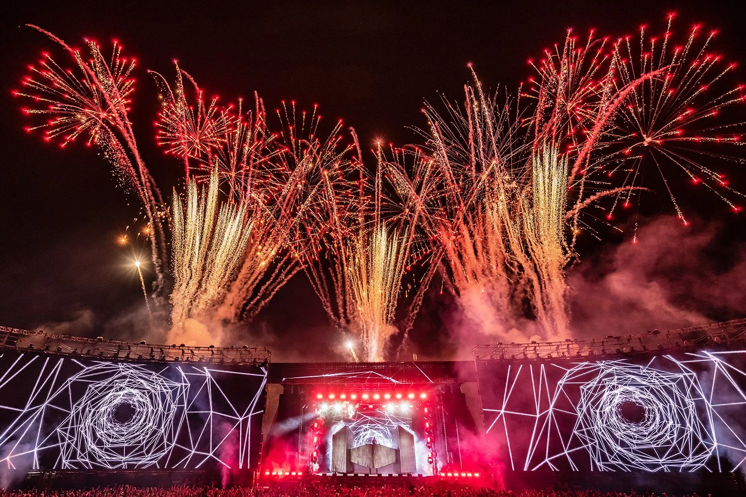 Fireworks display lighting up the night sky above a concert stage with digital geometric white designs.
