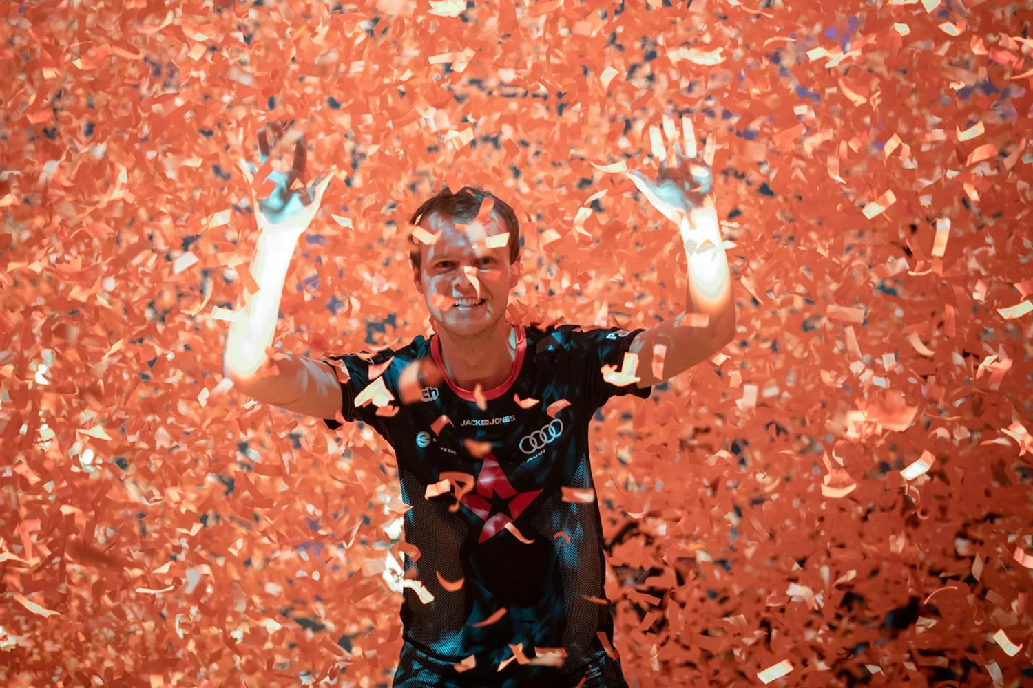 A man celebrating with a big smile, surrounded by flying orange confetti, wearing a black sports jersey with logos, during a moment of victory.