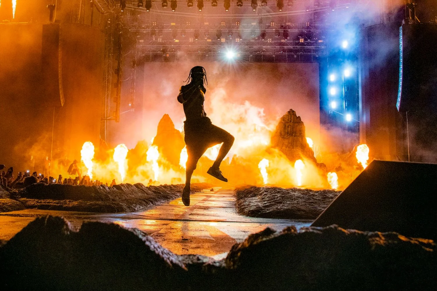A performer on stage, silhouetted against a fiery explosion and bright stage lights, in a concert or event setting.