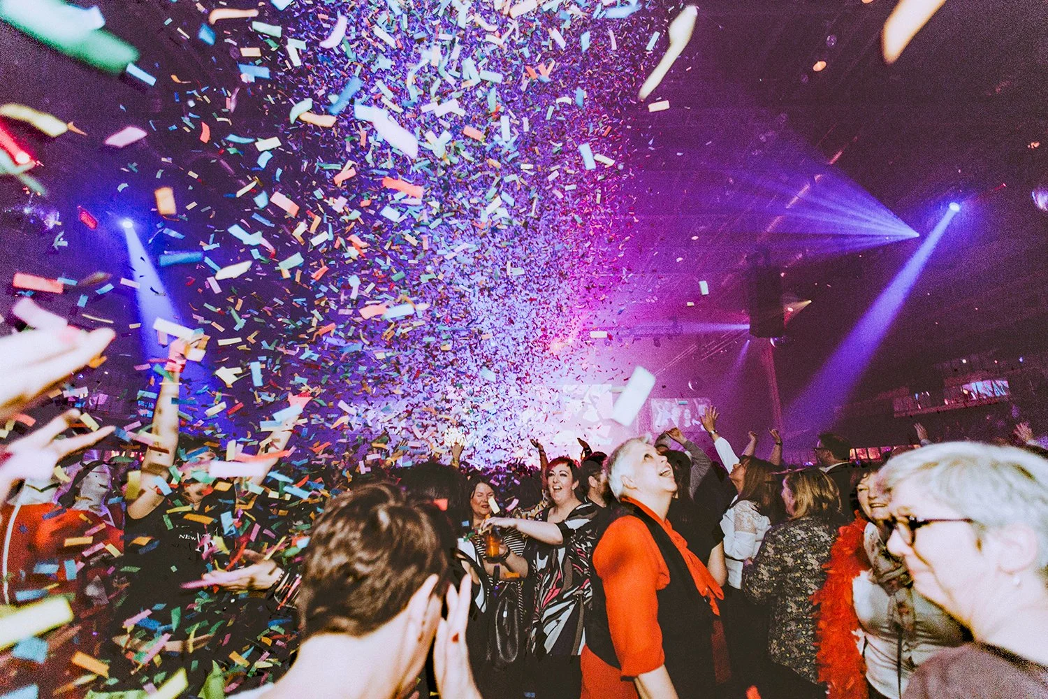 A lively party with colorful confetti falling from the ceiling, energetic crowd dancing and smiling, and vibrant purple and blue stage lights illuminating the scene.