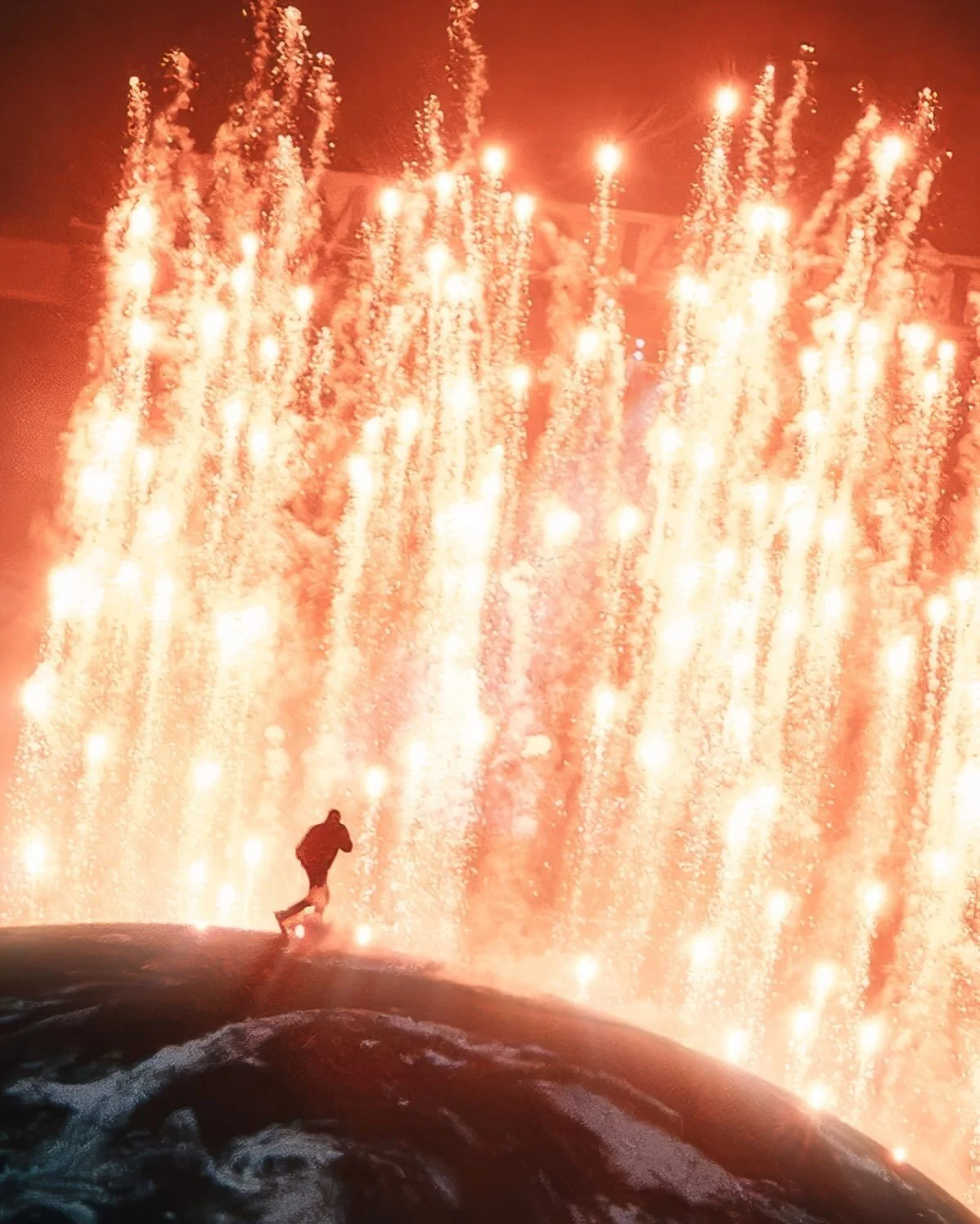 A person walking along a narrow log with waterfalls and sparks of fire in the background, illuminated by orange and yellow lights, creating a dramatic nighttime scene.