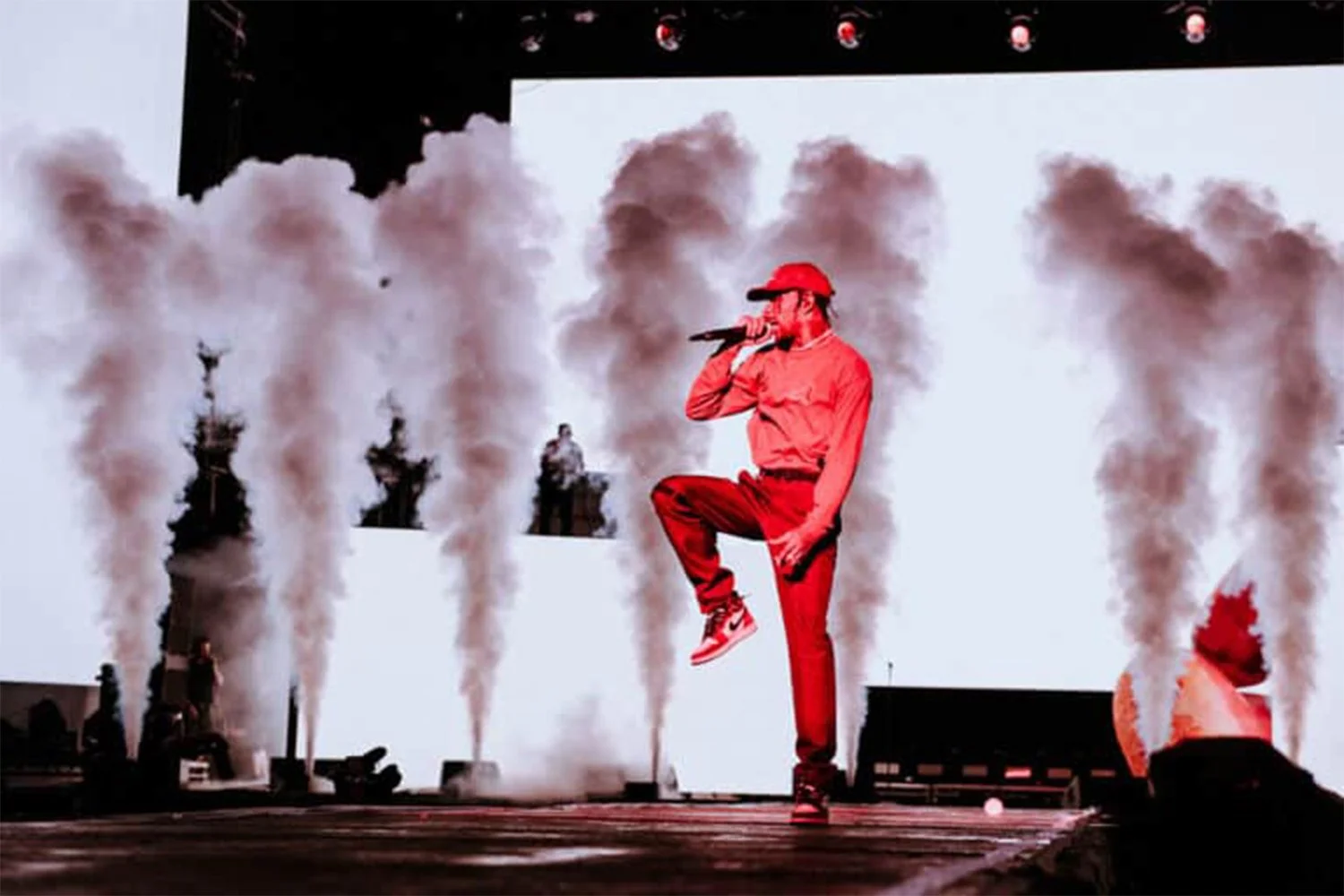 Performer dressed in red, wearing a baseball cap, singing into a microphone on stage with smoke effects behind him.