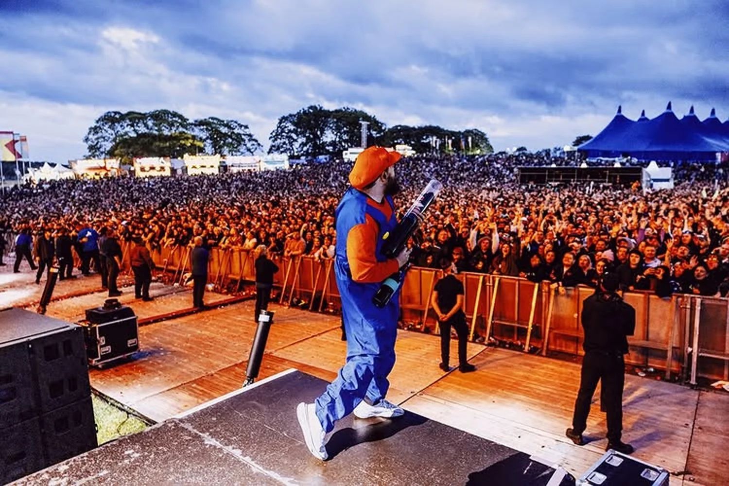 Performer dressed as Mario from video games on stage at an outdoor concert, holding a guitar, in front of a large audience under a cloudy sky.