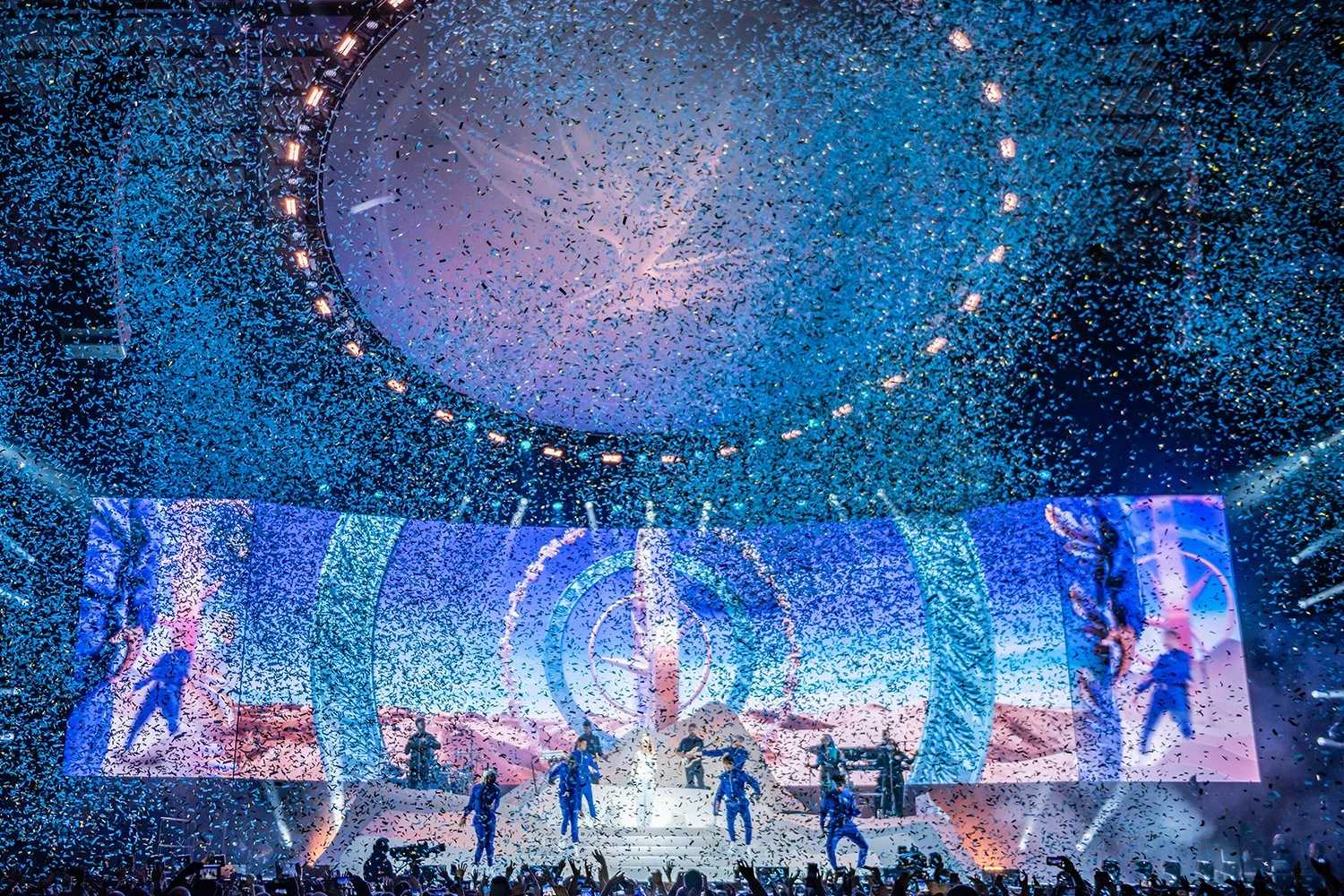 A concert stage illuminated with blue and purple lighting, dancers performing, and a large screen displaying visuals; confetti falling from above.
