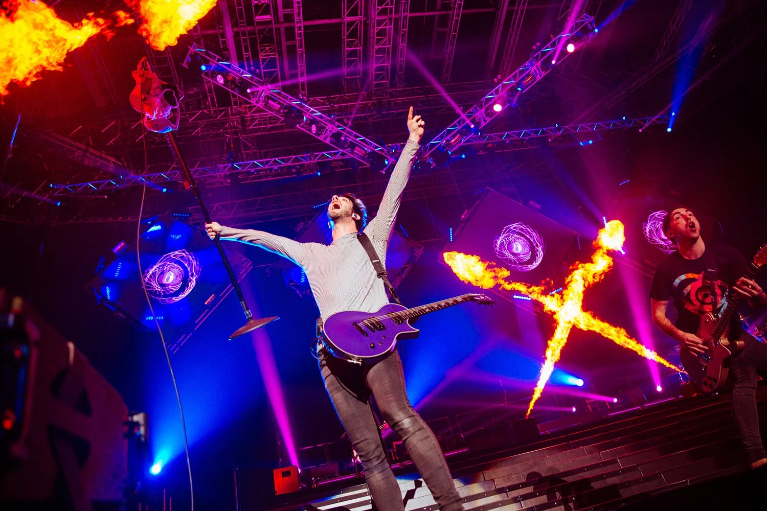 A live concert with two musicians performing on stage surrounded by vibrant purple, blue, and pink lighting. The main performer is playing an electric guitar, singing passionately, with arms raised. Fire and pyrotechnic effects are visible behind them, creating a dynamic and energetic atmosphere.