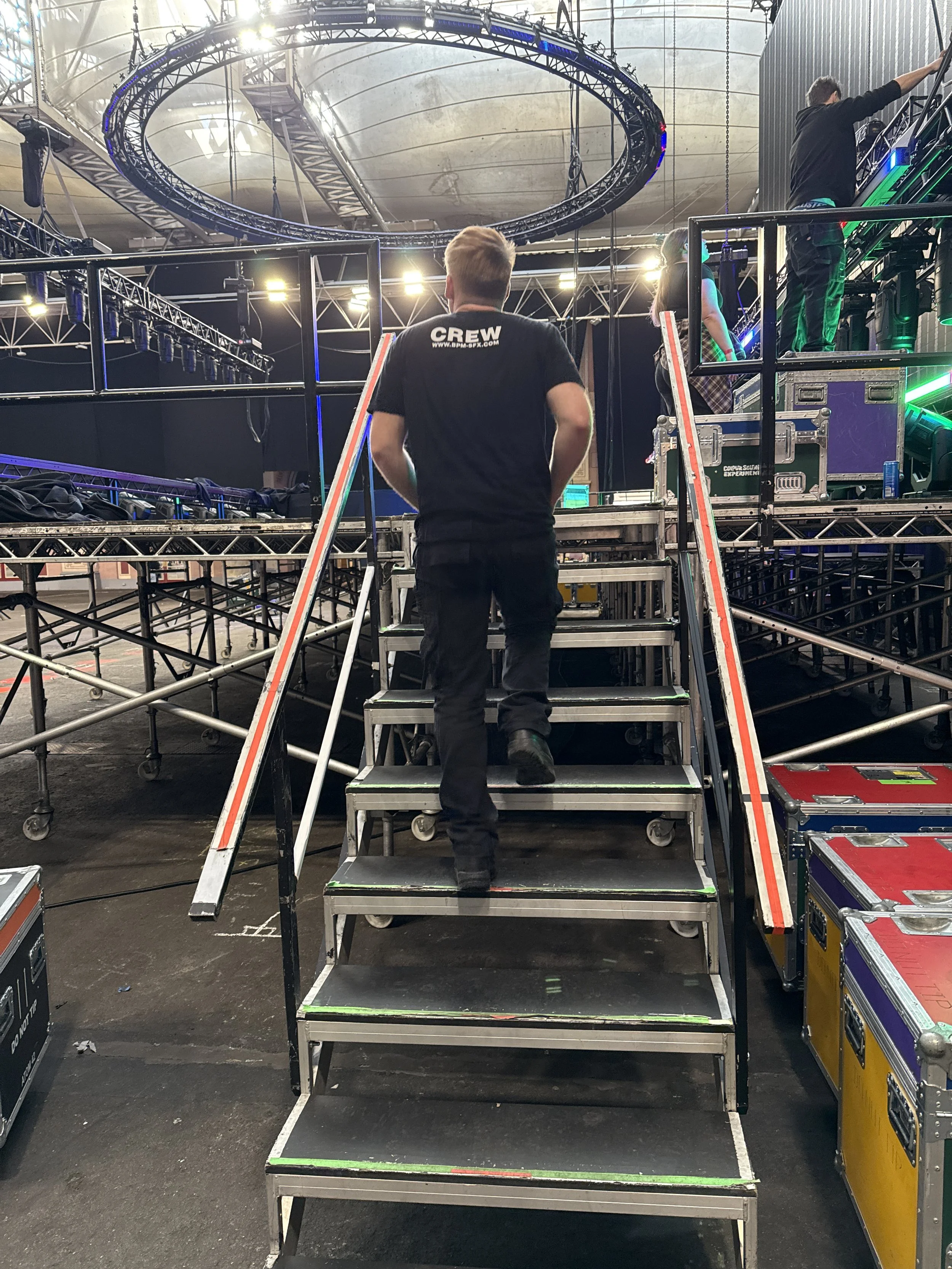 A worker is walking up a set of metal stairs in a large indoor event space, with stage lighting and rigging overhead, as other crew members prepare for an event.
