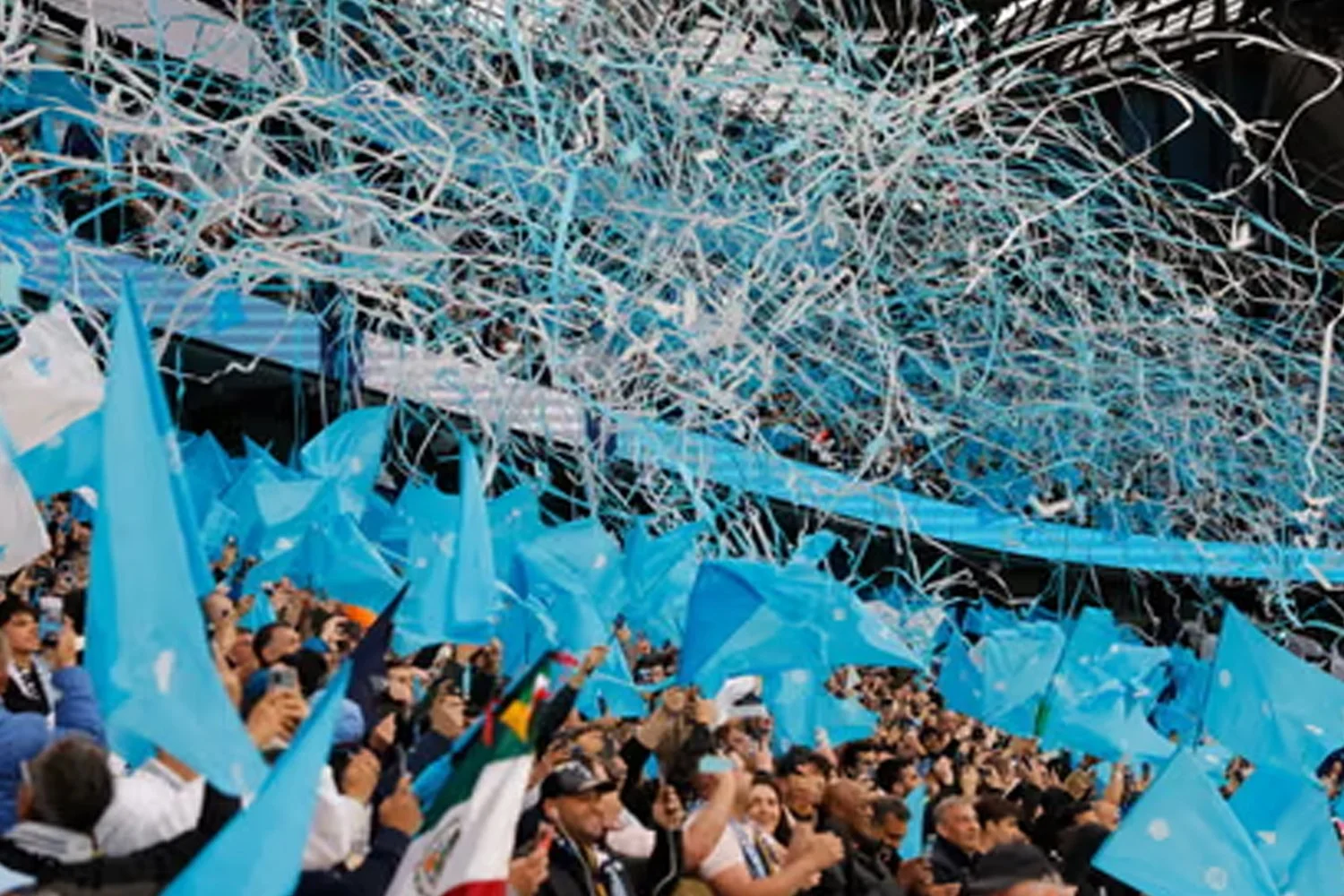 A large crowd of people holding blue flags at a stadium, with white and blue streamers overhead, likely during a sports event or celebration.