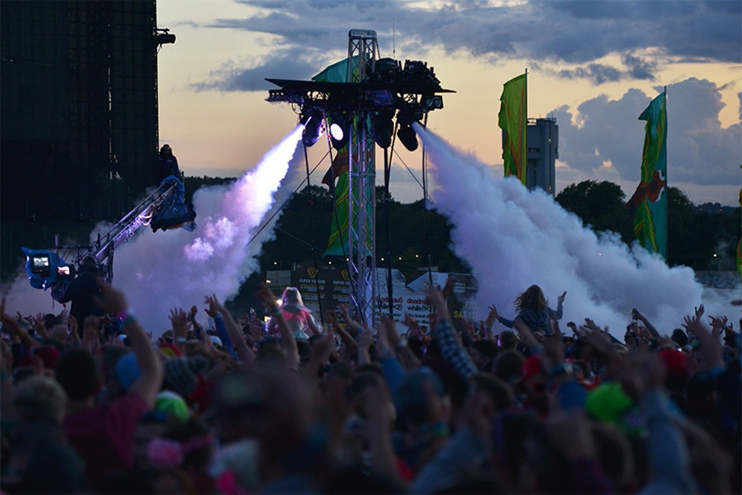 A concert or festival crowd watching a stage where colorful fireworks or pyrotechnics are being launched, with people raising their hands during the evening or sunset.